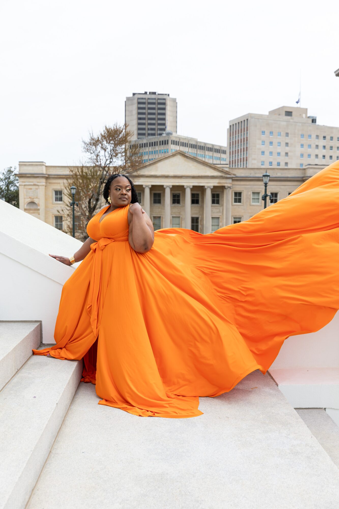 Woman in an orange dress sitting on stairs with a city building in the background.