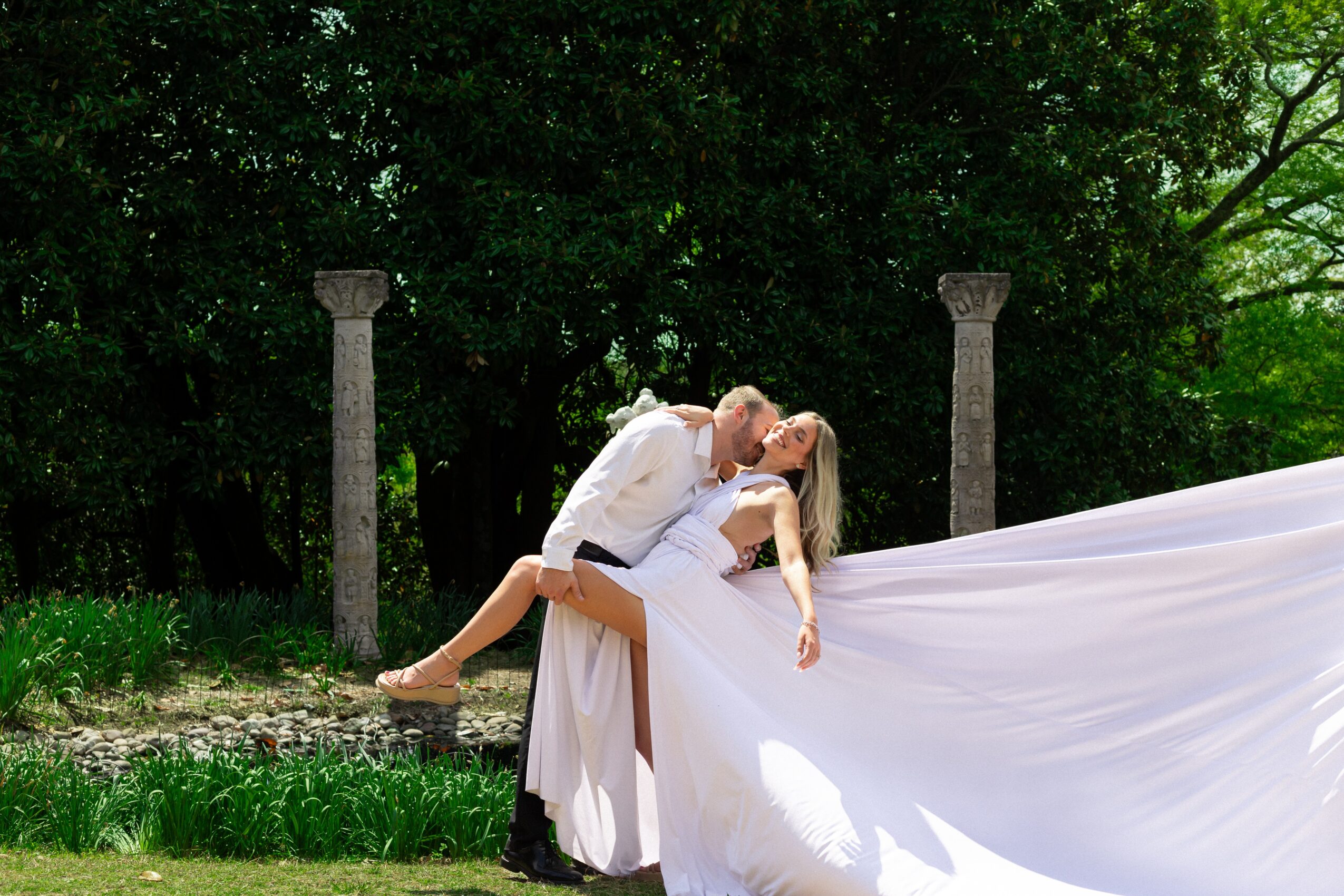 A couple in wedding attire sharing a kiss outdoors, with the groom holding the bride, her dress flowing behind her, green trees in background.