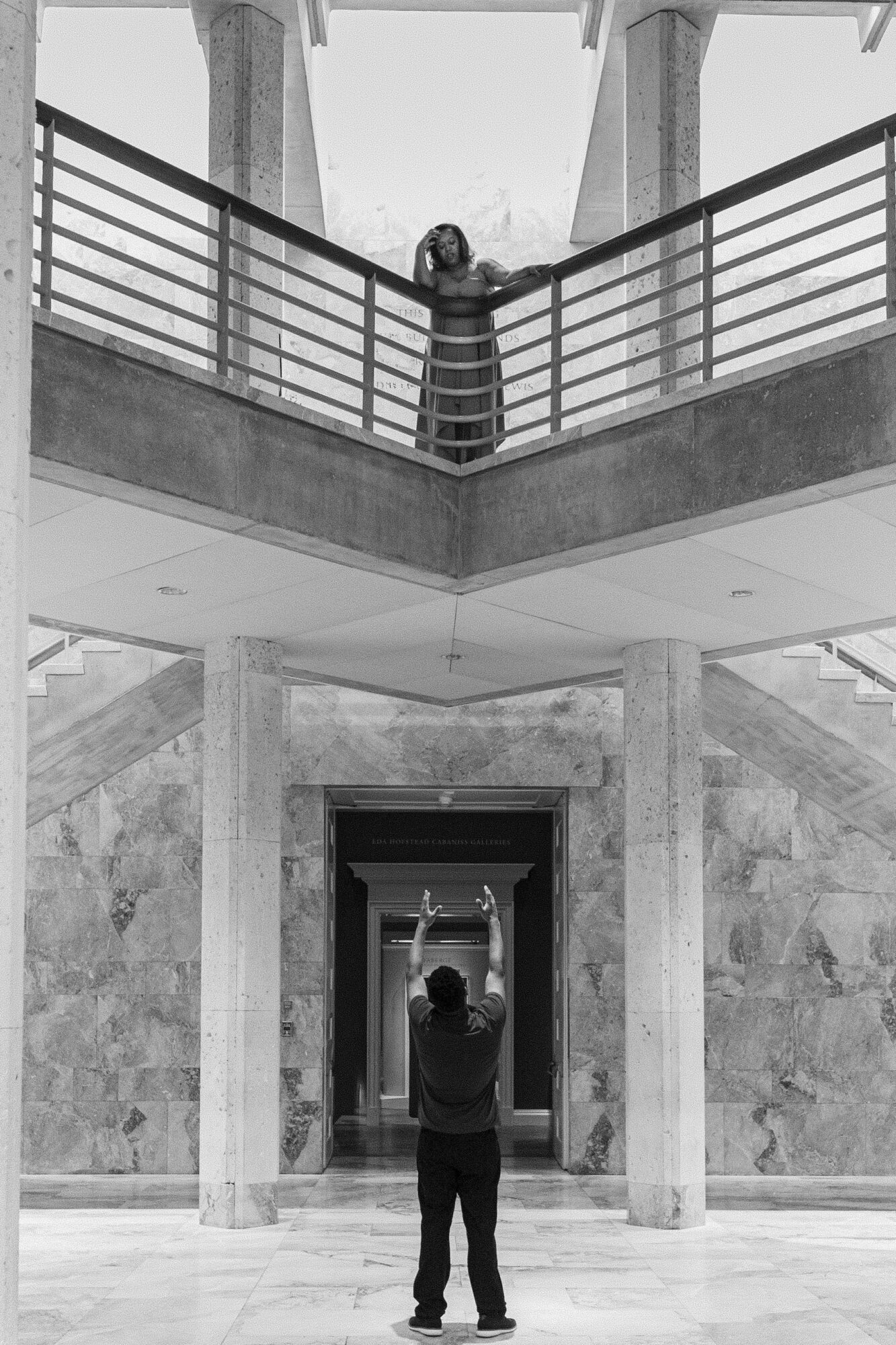 Person standing in a building with marble walls, looking up at another person on an upper balcony, arms raised.