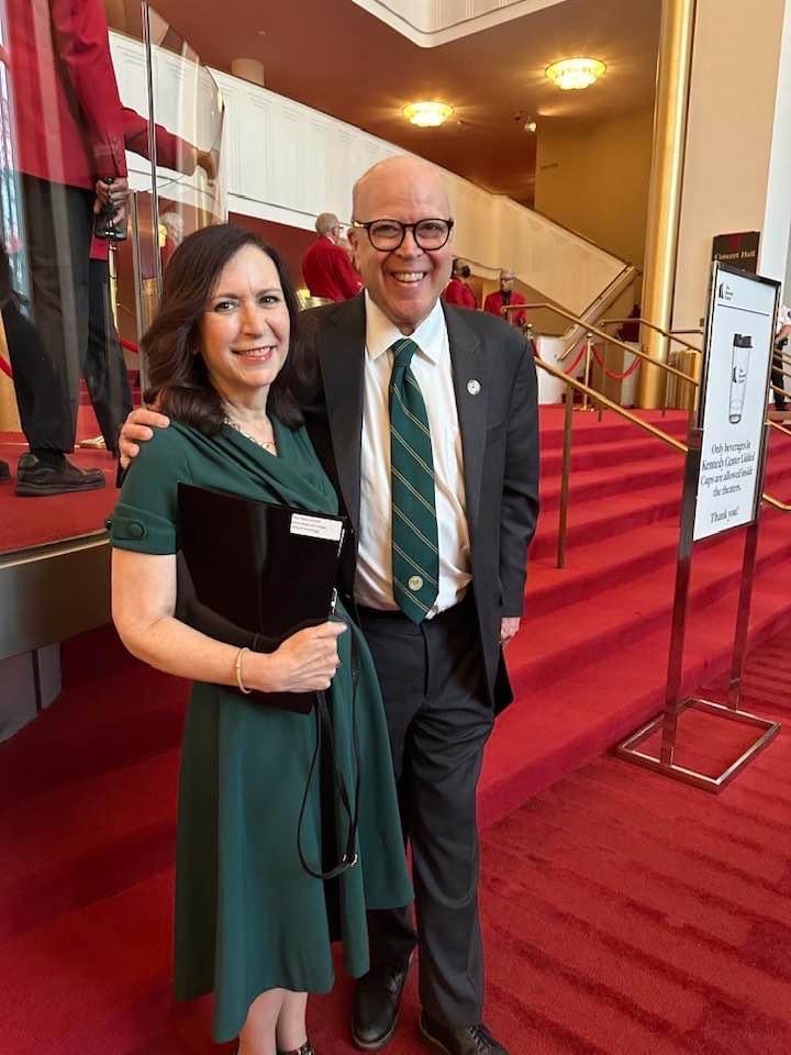 Two people, a woman and a man, stand together smiling in a red-carpeted venue with a staircase behind them.