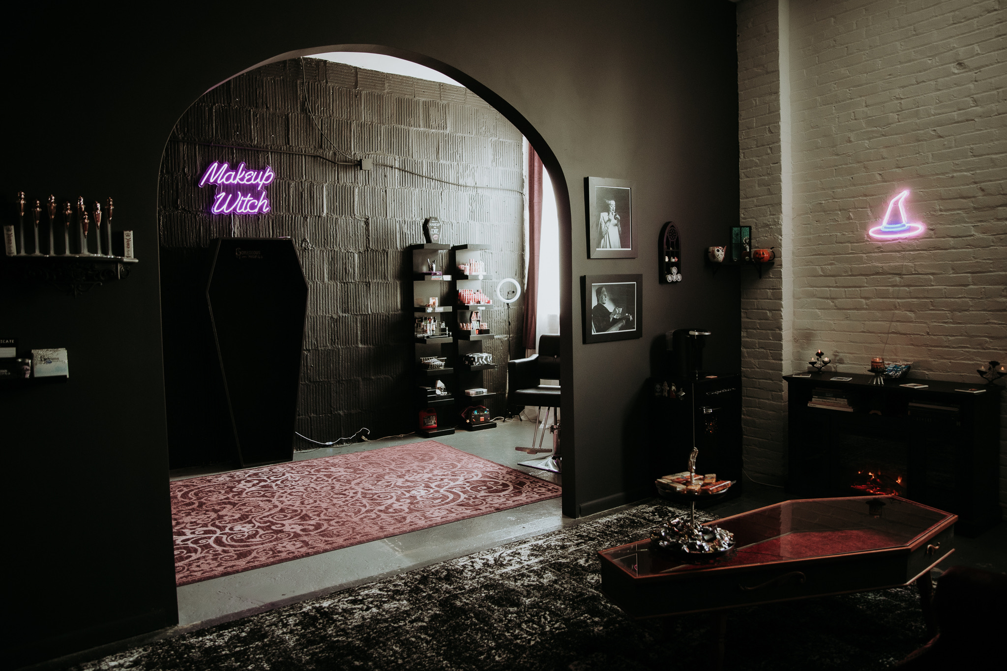 Interior of a makeup studio with a neon sign, shelves, framed pictures, and a seating area, viewed through an archway.