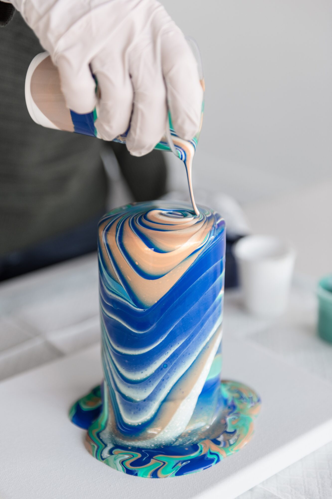 Person wearing a glove pours colorful liquid onto a tall, cylindrical object with blue, white, and gold swirls.