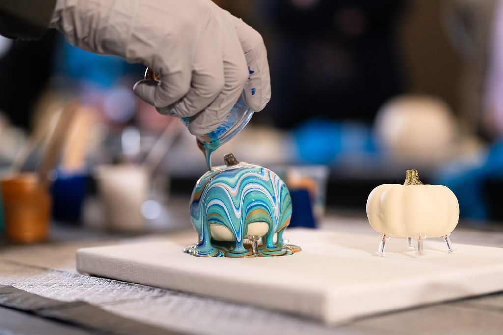 Person decorating a pumpkin-shaped object with blue and white swirls, on a white surface in a workshop setting.