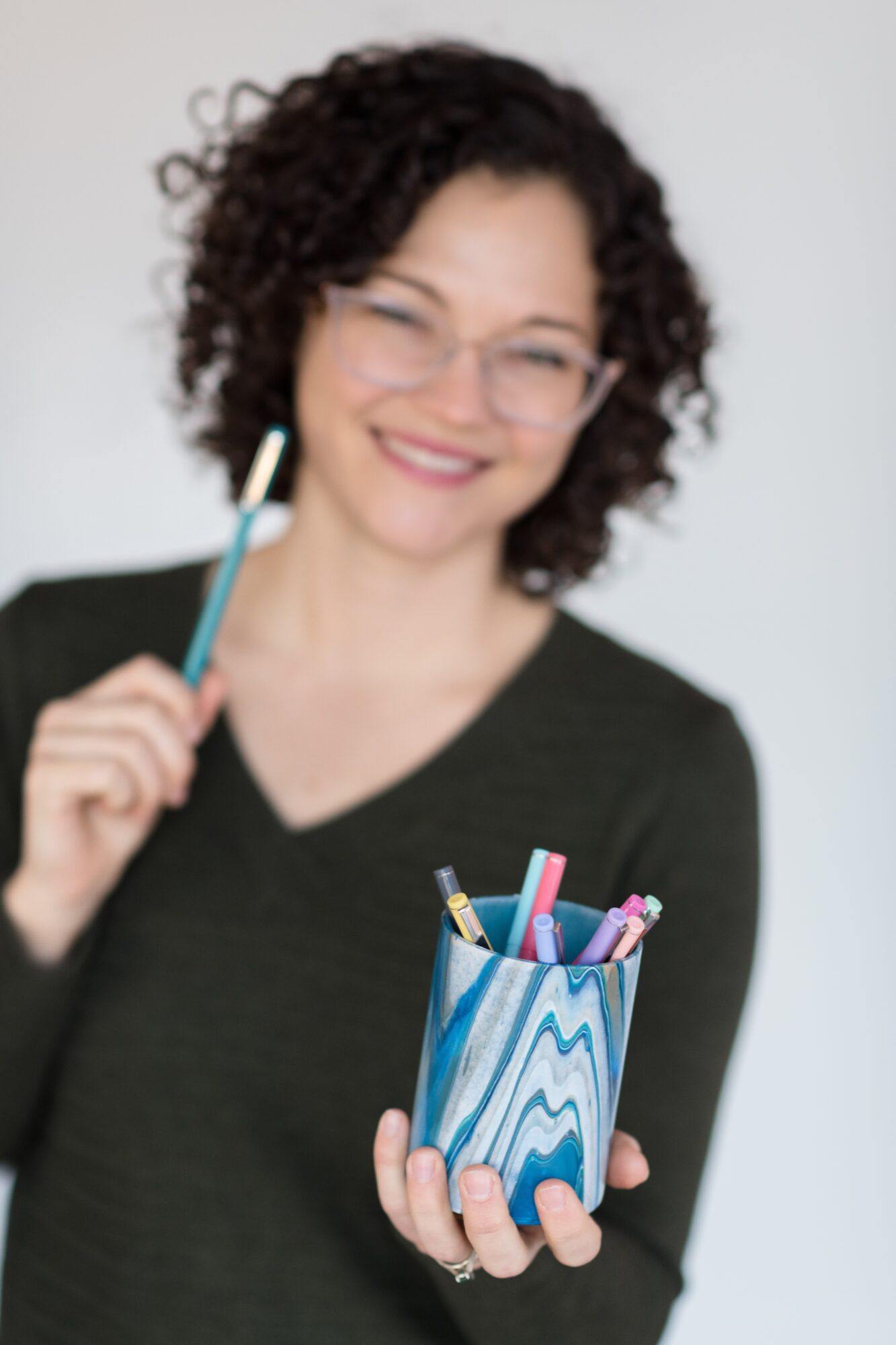 Woman with curly hair and glasses holding a pencil, smiling, with a container of colorful pens and pencils, against a plain background.