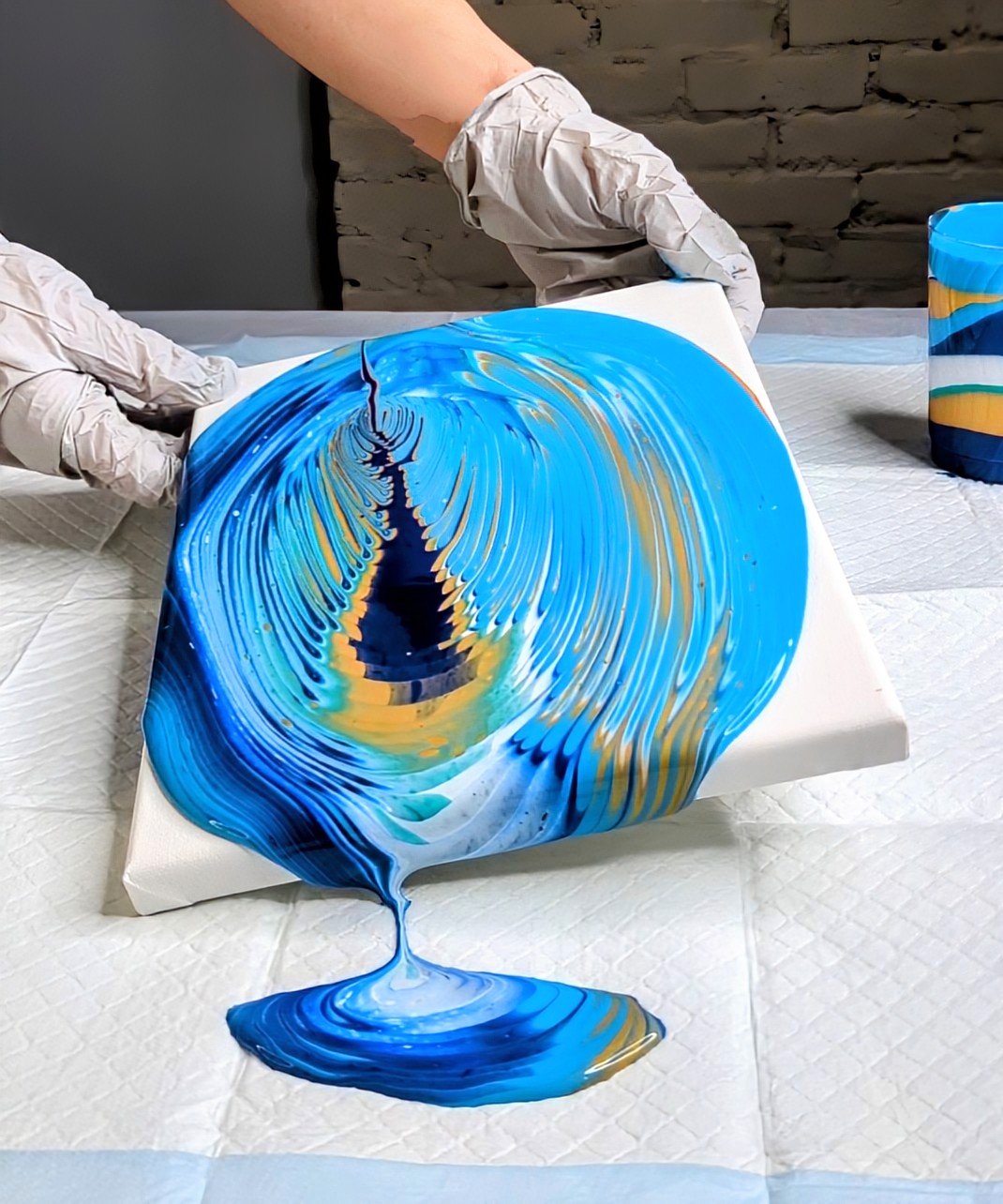 Person wearing gloves holding a canvas with blue, yellow, and black paint swirling, dripping onto a surface.
