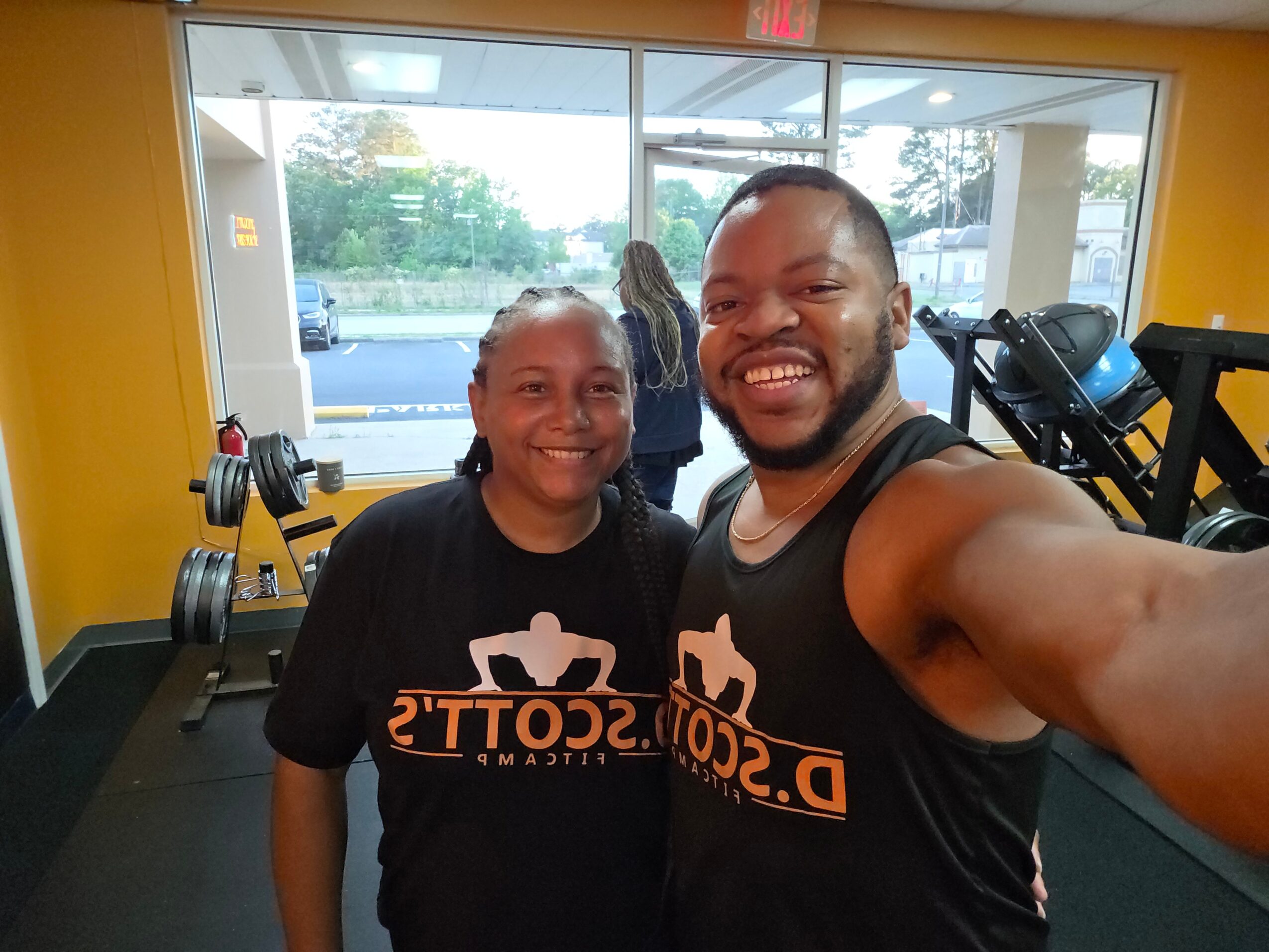 Two smiling people in a gym, wearing black shirts with orange text, taking a selfie with gym equipment in background.
