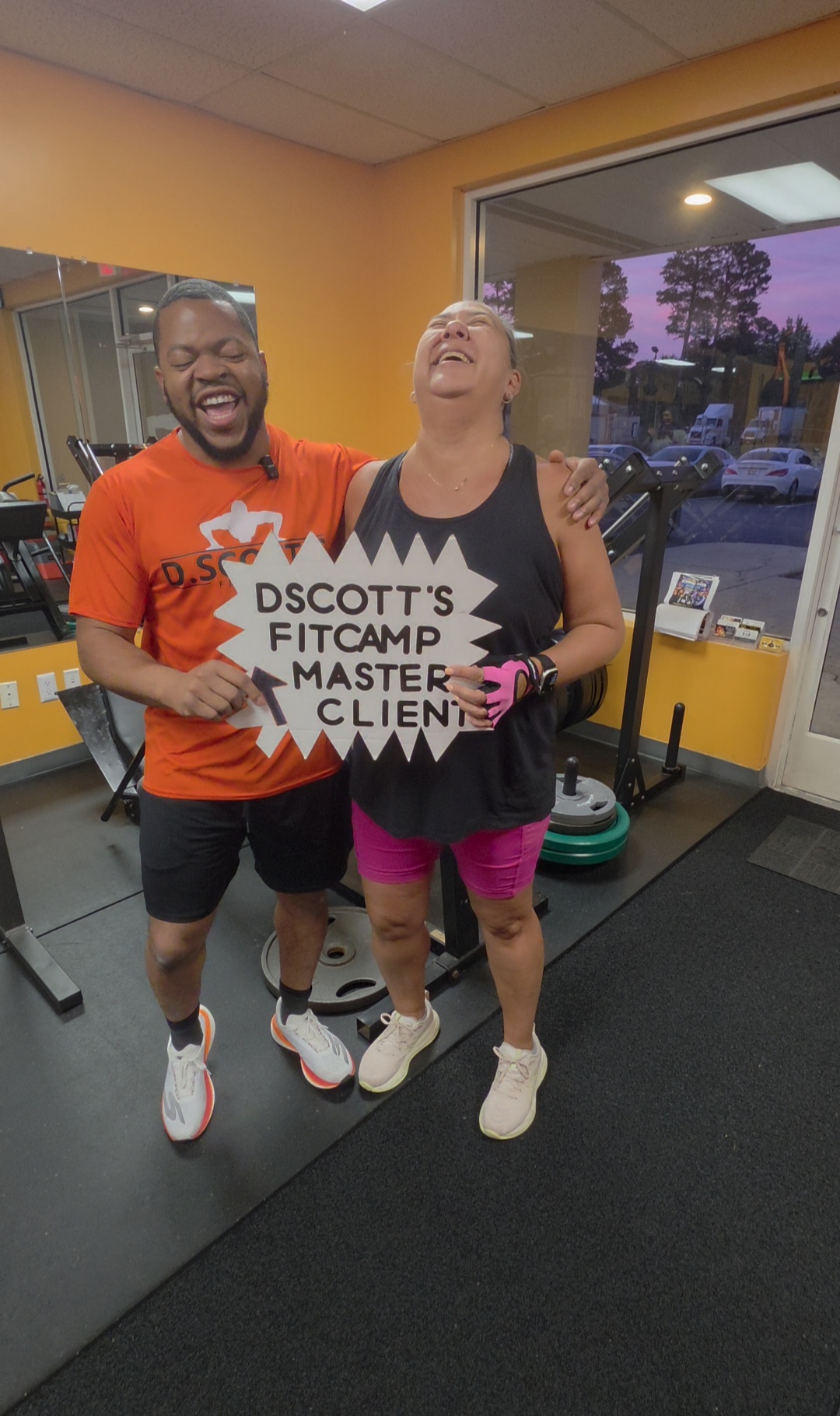 Two people smiling and hugging inside a gym, one holding a sign, with gym equipment and windows showing trees outside.