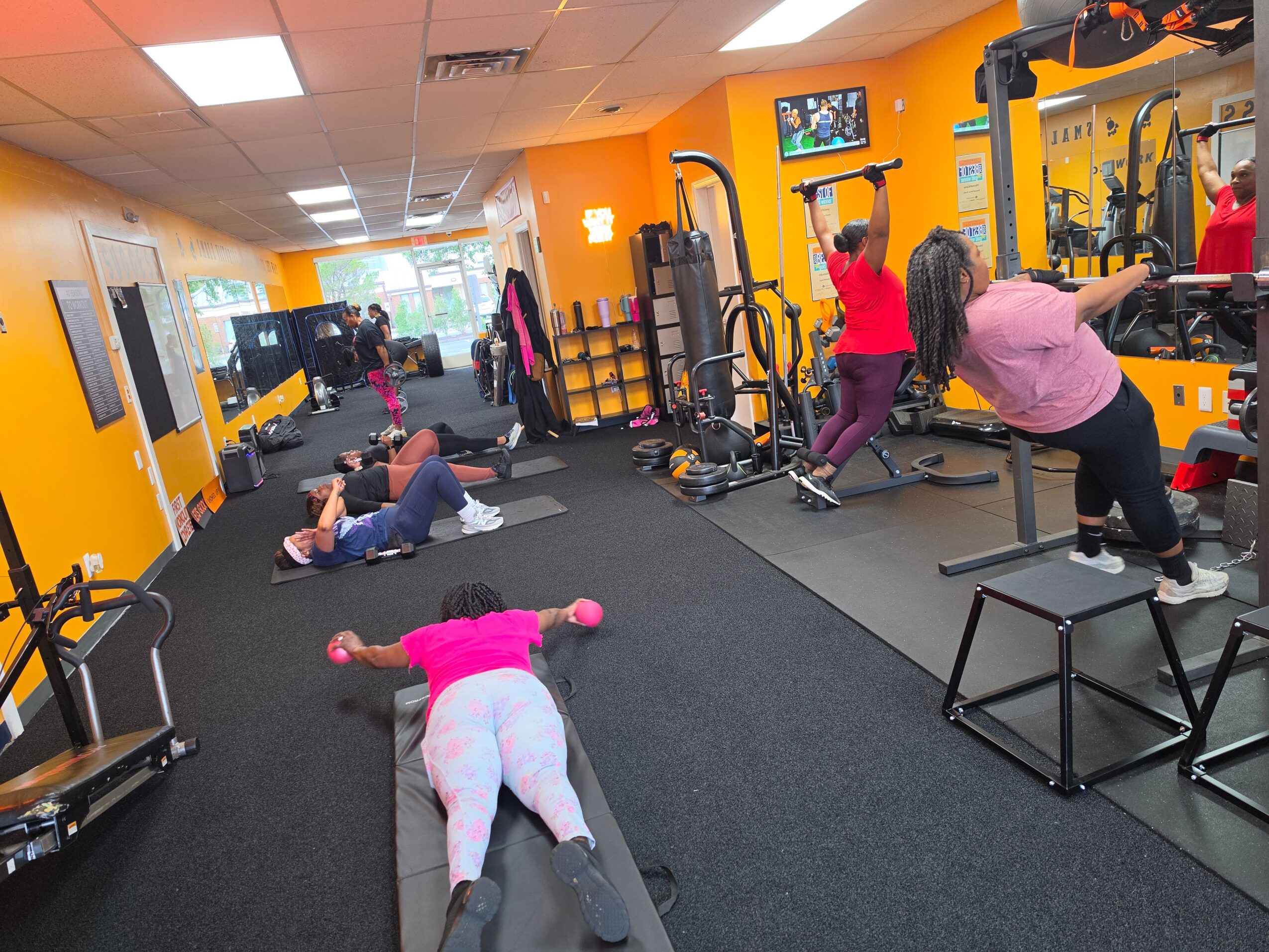 People exercising in a brightly lit gym with yellow walls, weight machines, and mats on the floor.