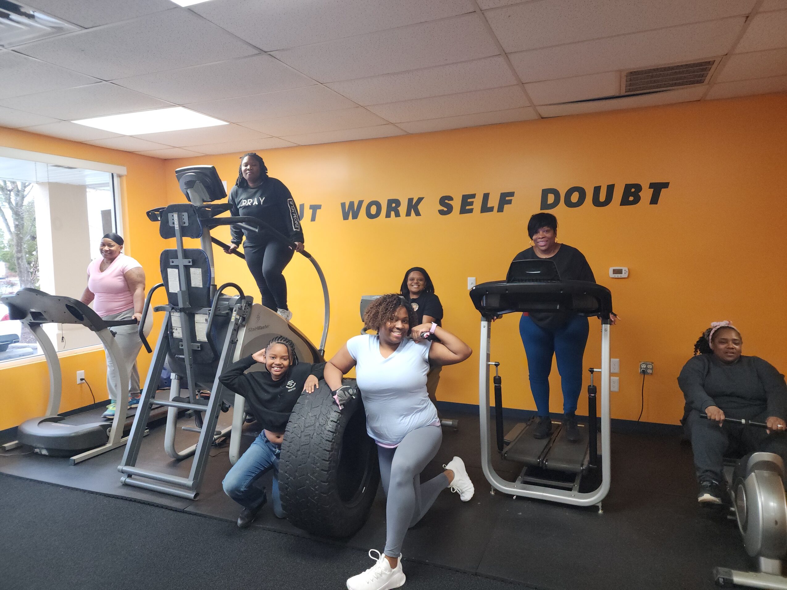 Group of people exercising in a gym with yellow walls and motivational words, including a person on a treadmill and others using equipment.