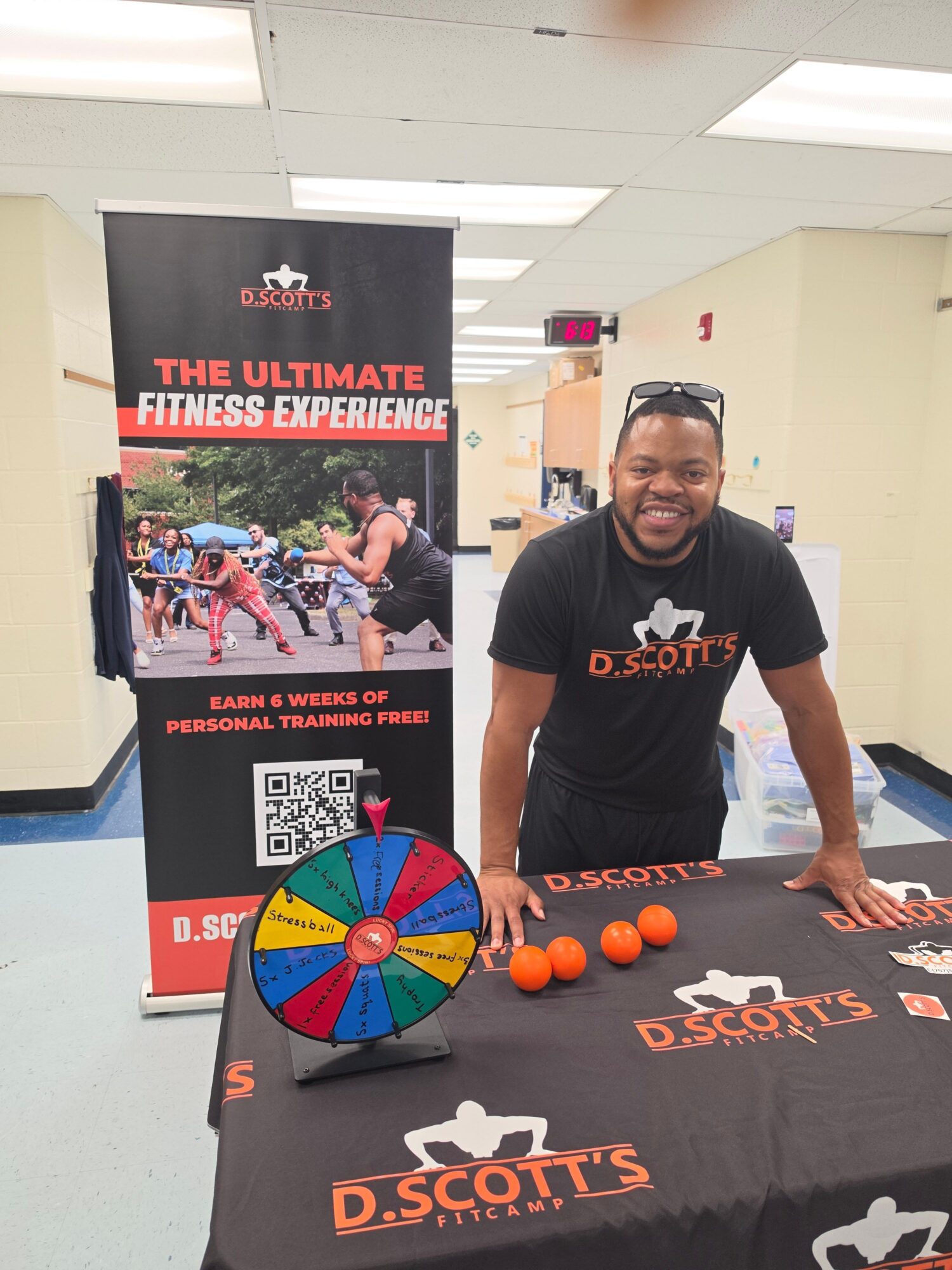 Person standing at a table with orange balls and a colorful wheel, indoor setting, promotional banners in background.