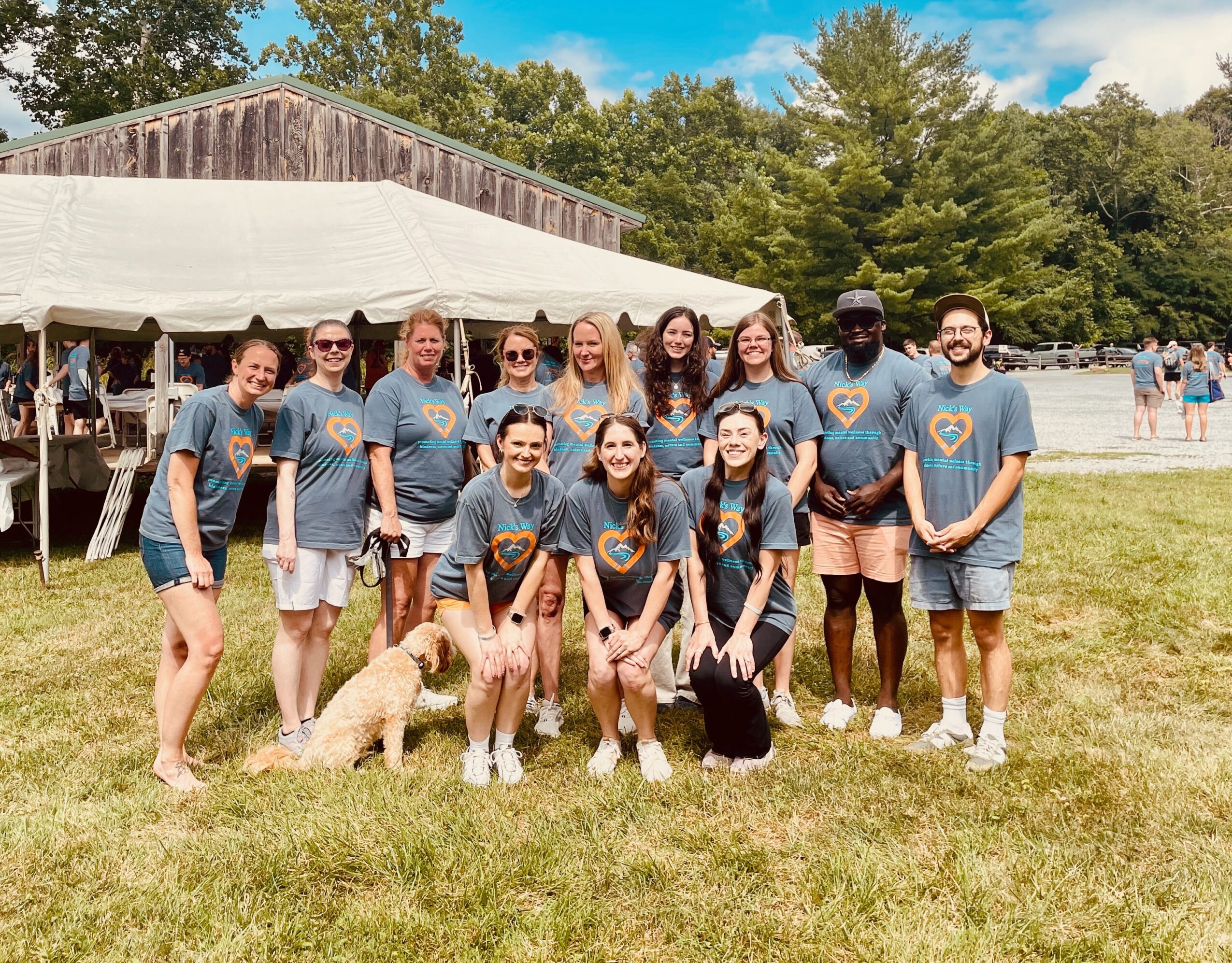 Group of people in matching shirts standing on grass with trees and a tent in background.