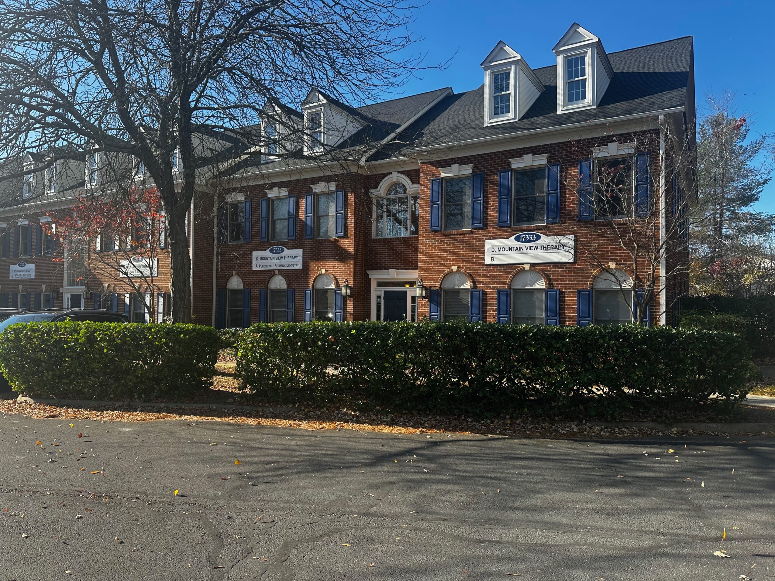 Three-story brick building with dormer windows, surrounded by trees and a hedge, under a clear blue sky.