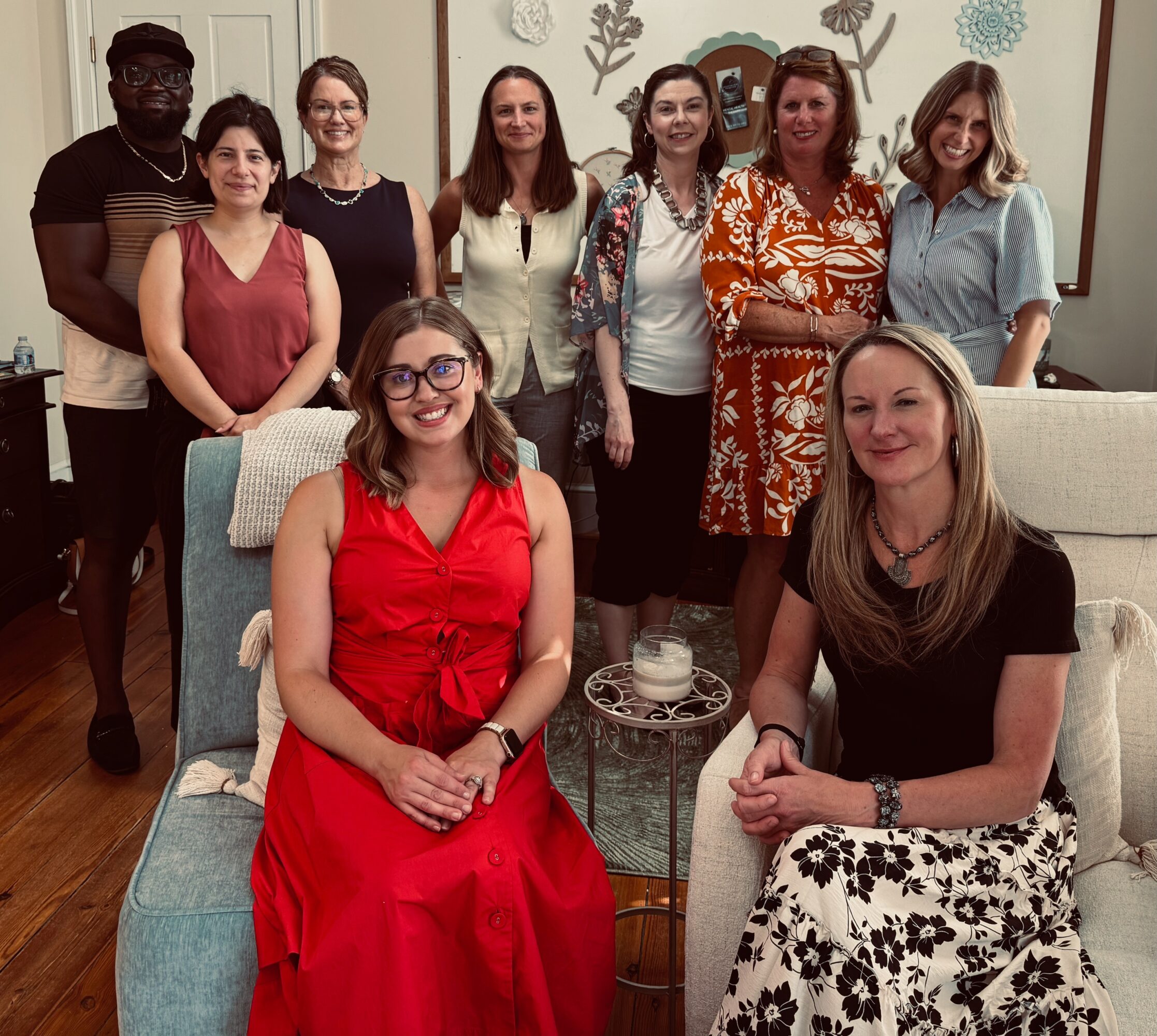 Group of nine women and one man gathered in a living room, some sitting and some standing, smiling at the camera.