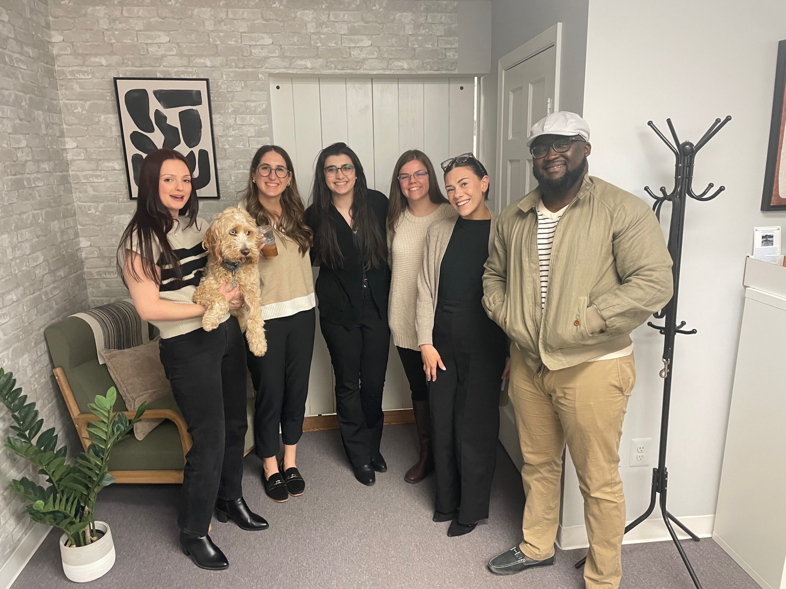 Group of six people standing indoors, one holding a small dog, with a plant and coat rack nearby.