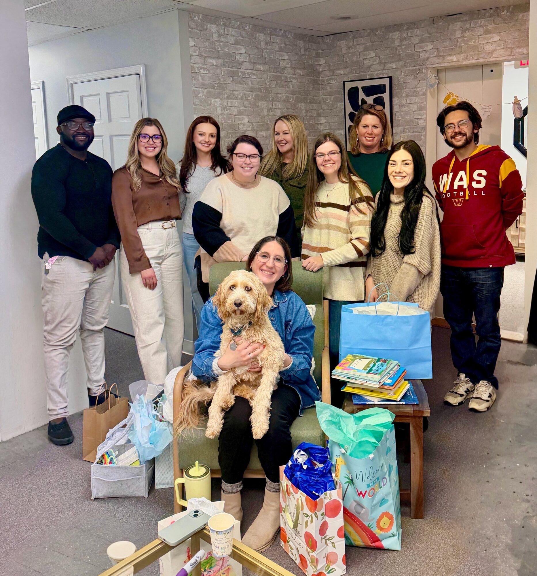 Group of ten people, including a woman with a dog, standing indoors with gift bags and boxes, smiling at the camera.