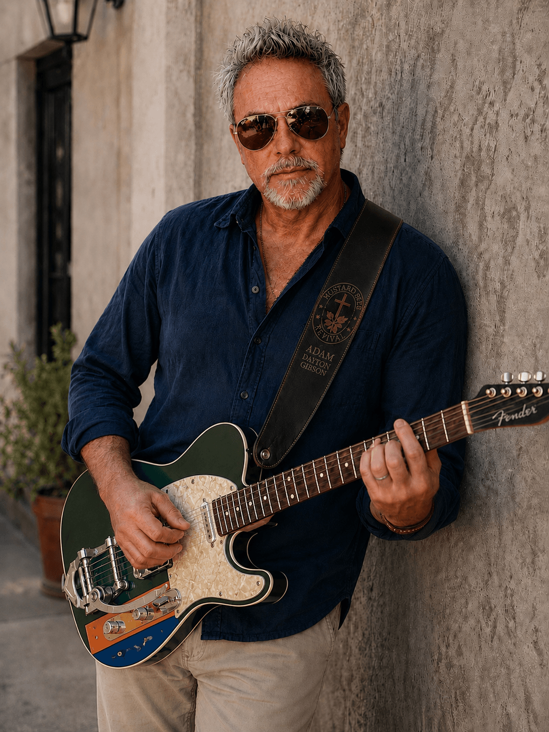 Man with sunglasses playing electric guitar against a textured wall, wearing a dark shirt and light pants.
