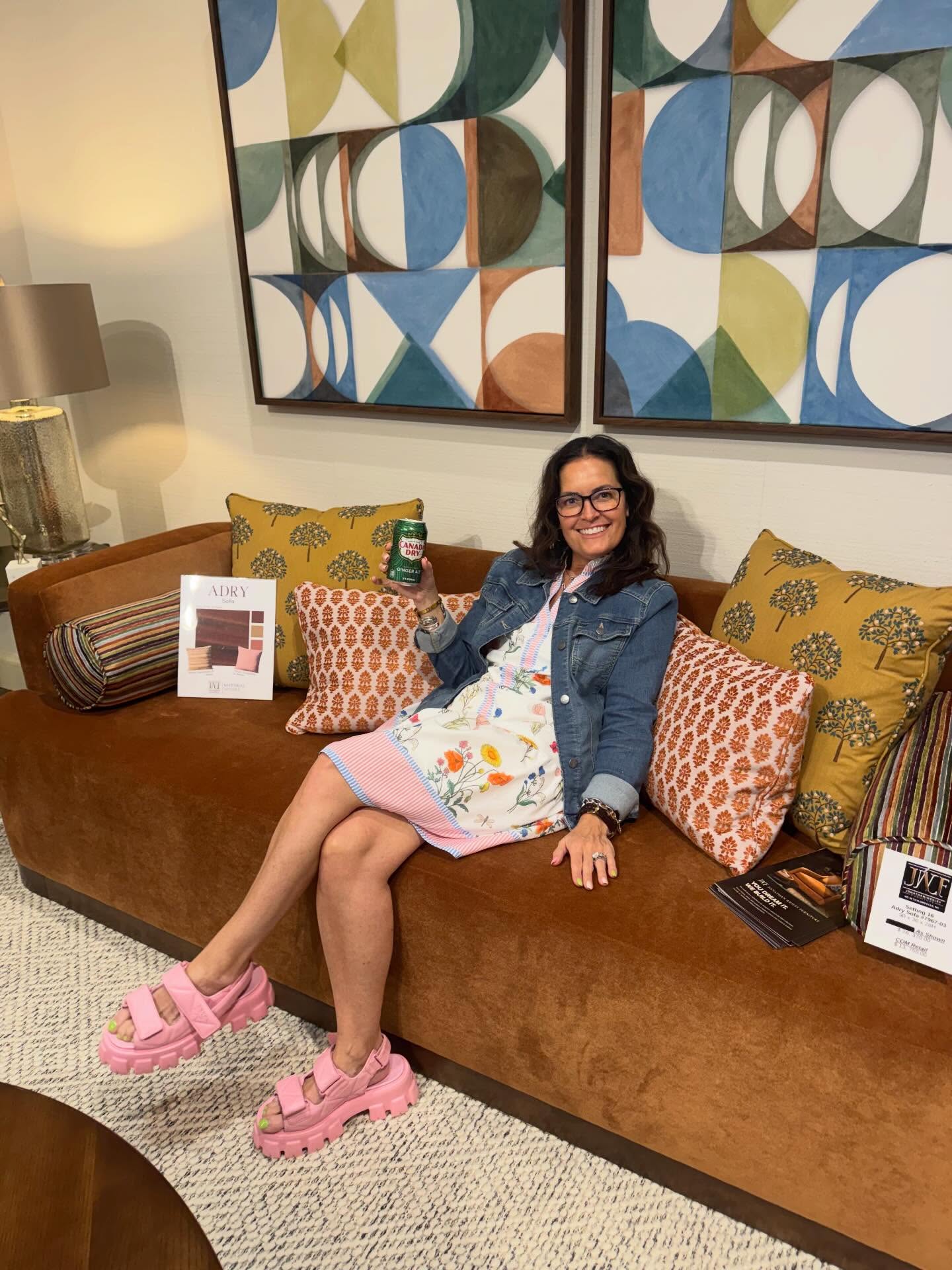 Woman sitting on a brown sofa with colorful pillows, smiling, in a living room with abstract wall art.
