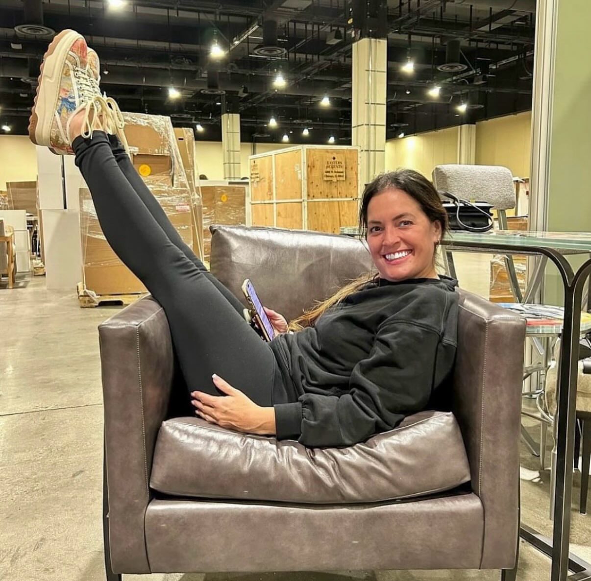 Woman sitting on a chair with legs raised, smiling, in a warehouse setting, holding a phone.