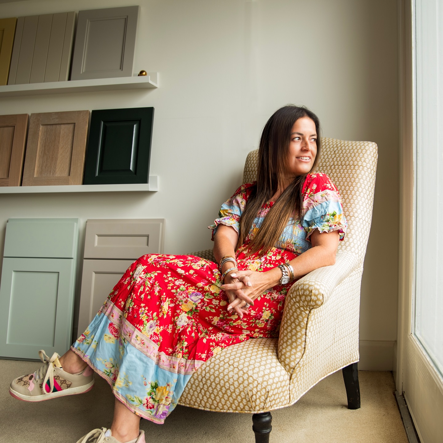 Woman sitting in a beige armchair near a window, wearing a colorful dress, smiling, with shelves of cabinet doors behind her.