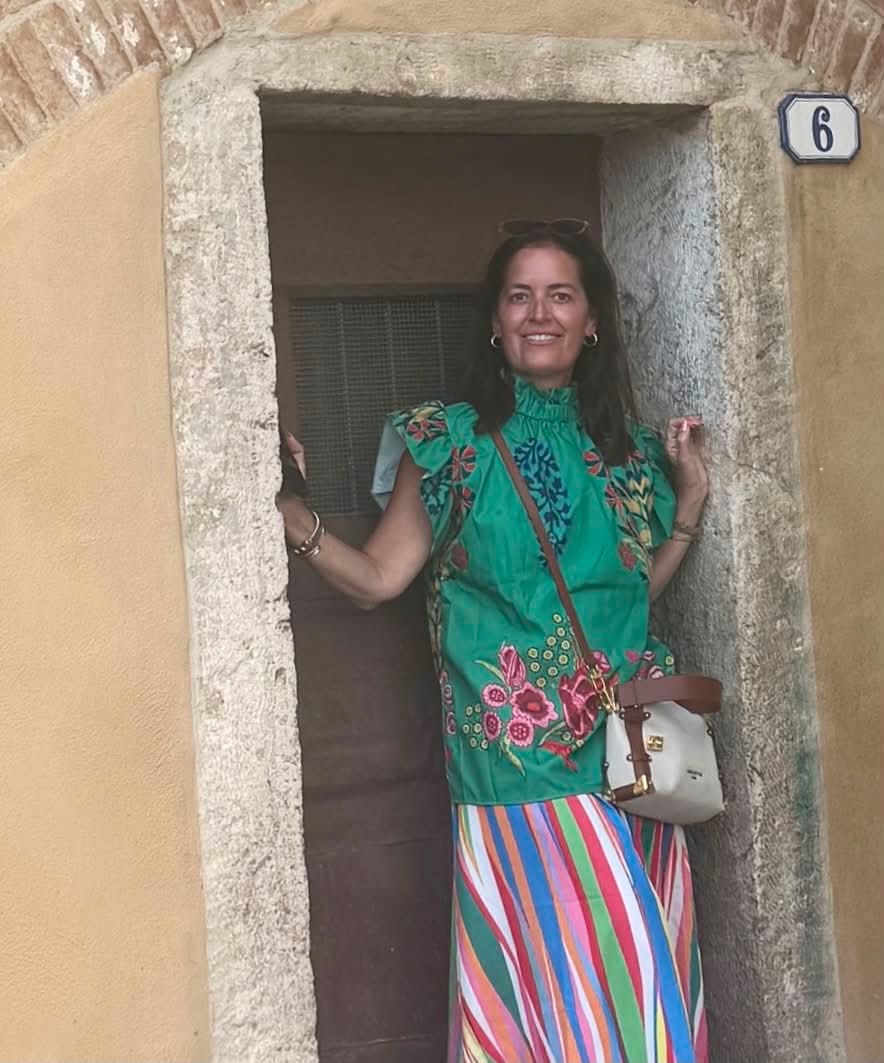 Woman standing in doorway, smiling, wearing a colorful striped skirt and green top with pink floral patterns, holding a bag.
