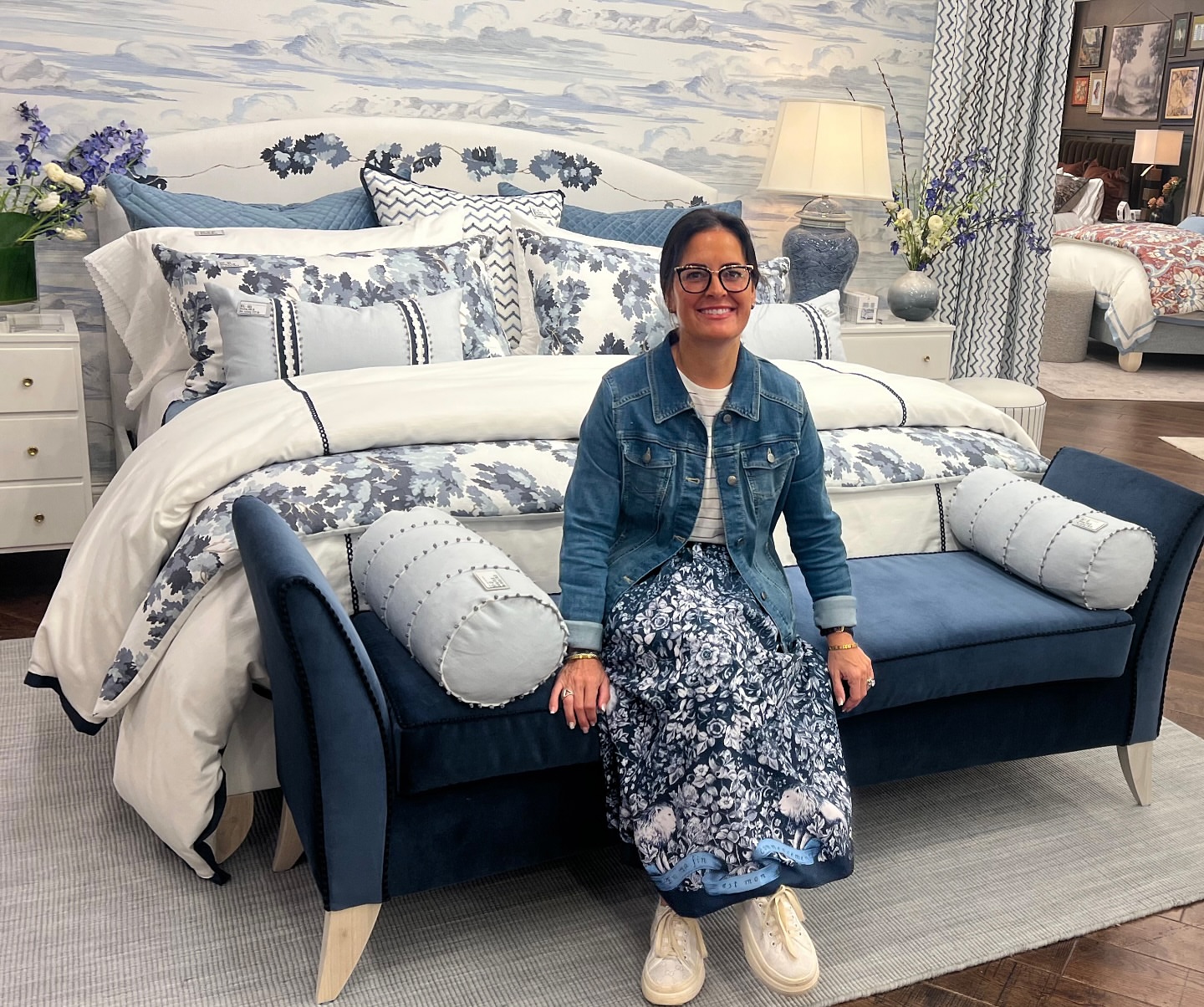 Person sitting on a blue sofa in a bedroom display with a bed, pillows, and floral decor, smiling at the camera.