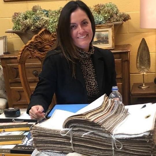Woman with dark hair smiling, sitting at a desk with a large stack of fabric samples, in a warmly decorated room.