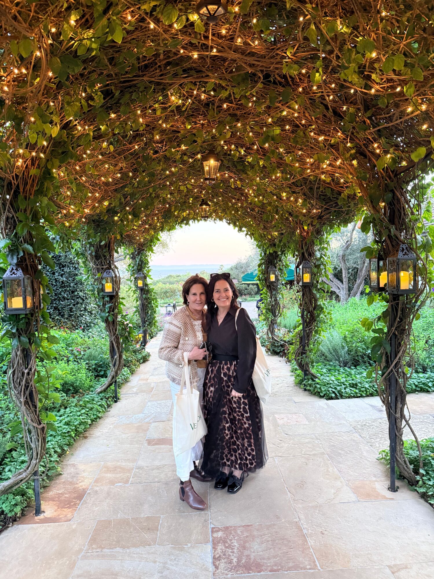 Two women walking along a garden path with trees and string lights overhead.