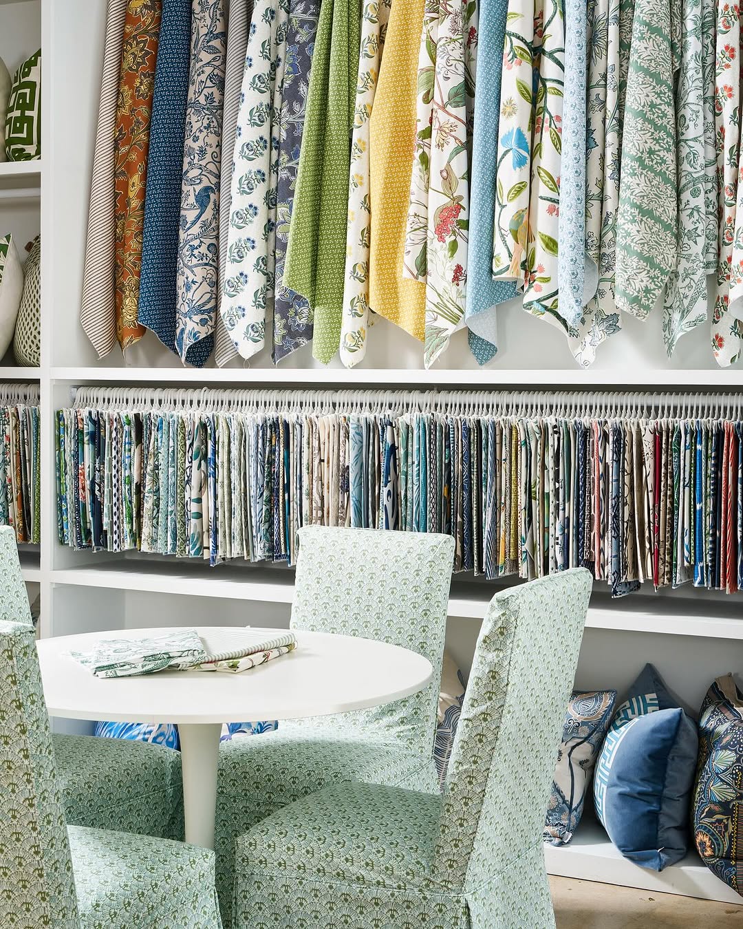 Clothing fabric samples hanging above a table with two chairs, fabric swatches on the table, and shelves with fabric rolls and bags.