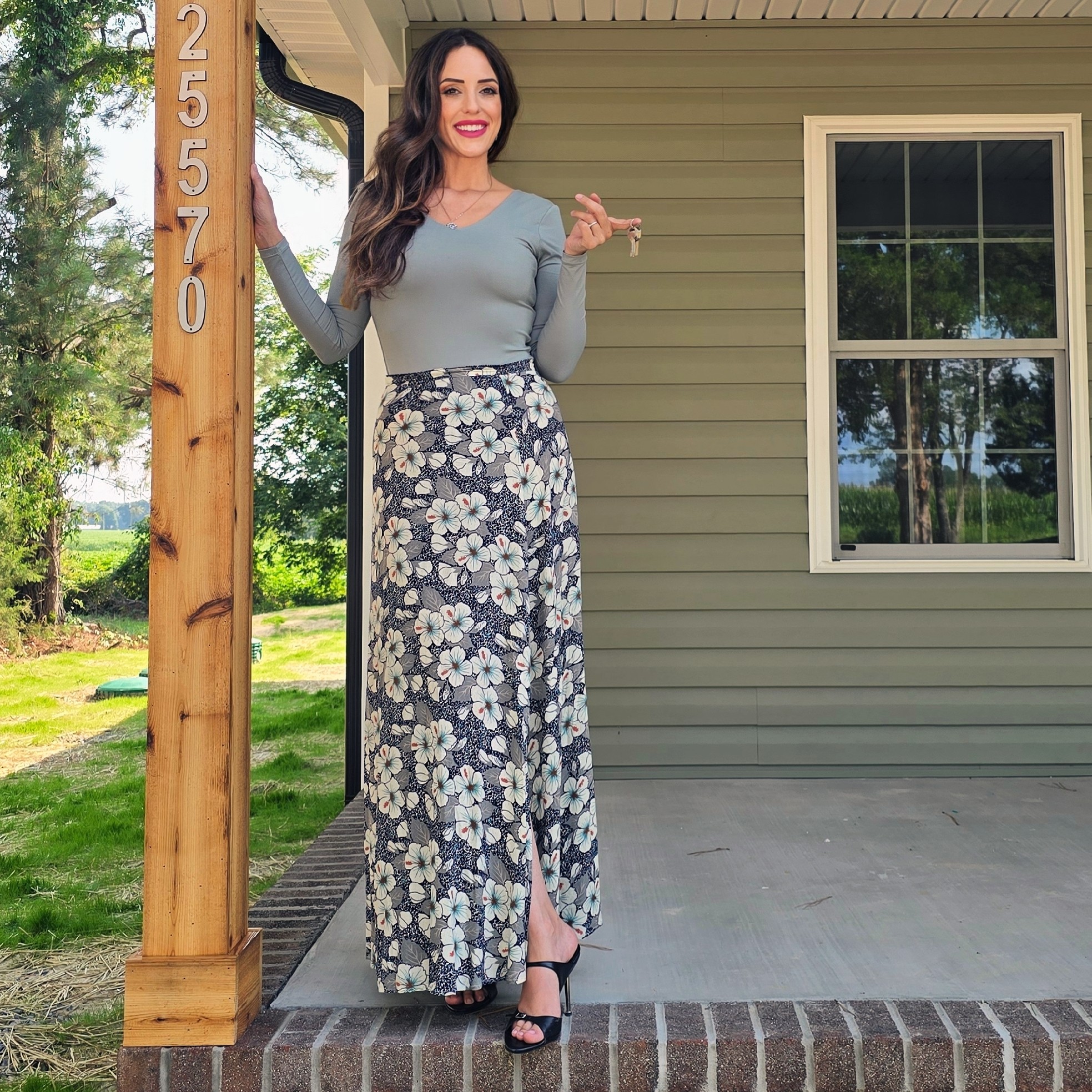 Woman standing outside next to a house with a window, wearing a long floral skirt and a light-colored top, smiling.