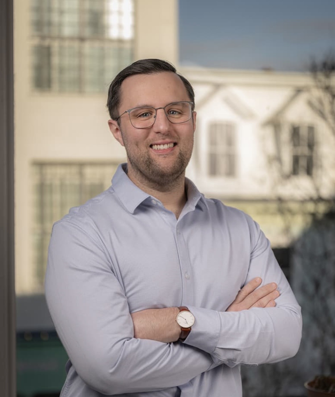 Man with glasses and a beard, smiling, wearing a light blue shirt, standing outdoors with buildings in background.