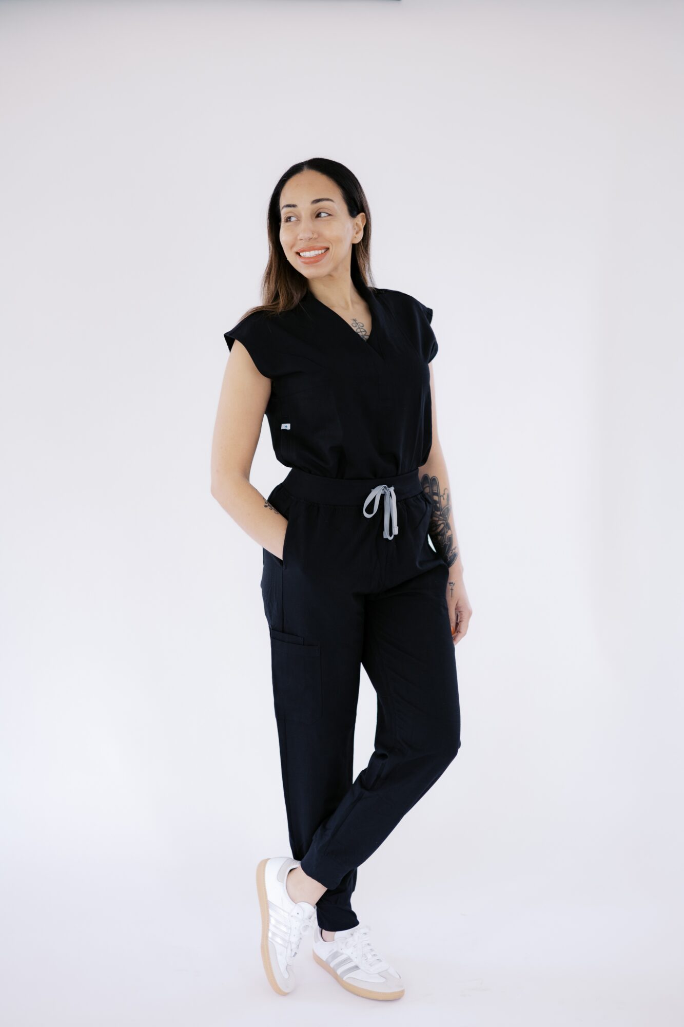 Woman standing with hands in pockets, smiling, wearing black top, black pants, and white sneakers, against a plain white background.