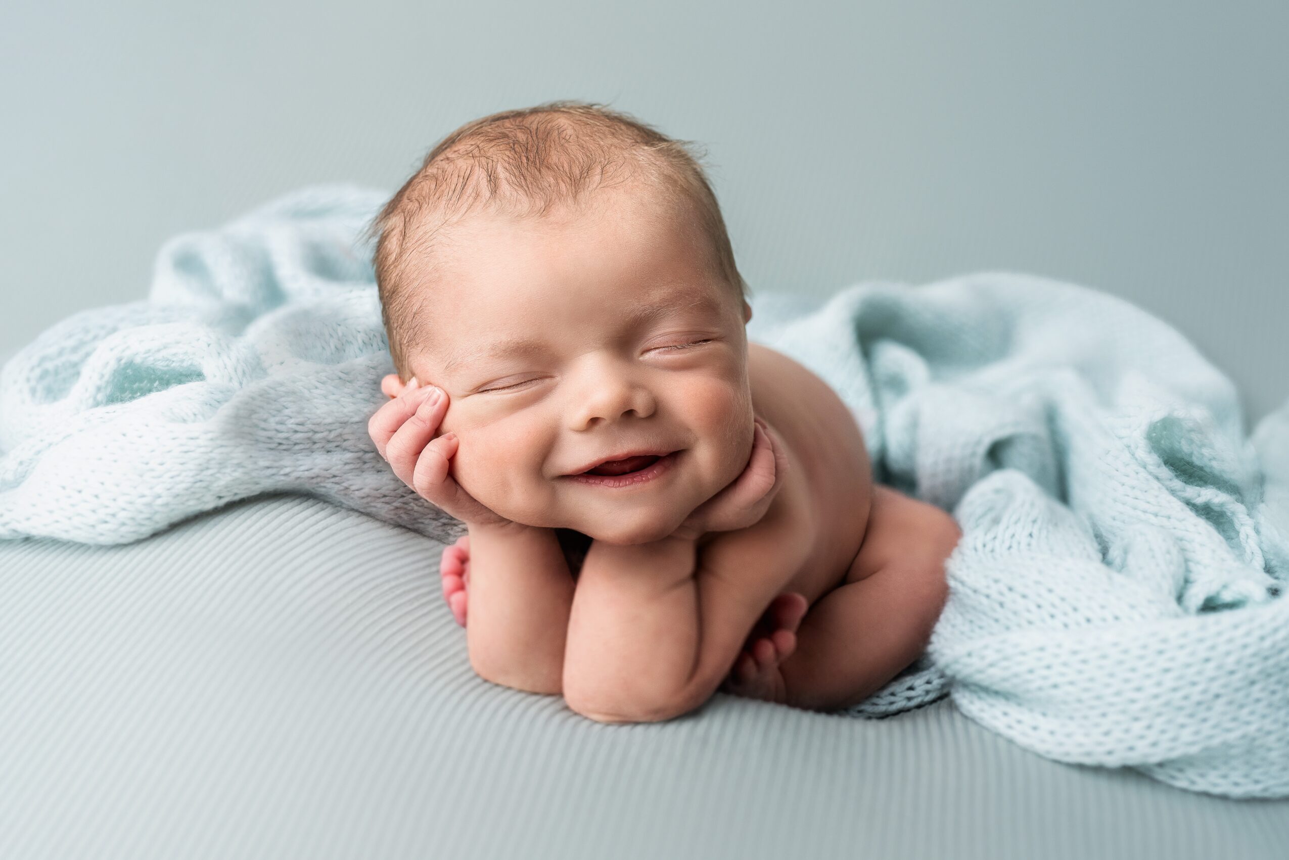 Smiling newborn baby lying on a soft surface, resting head on hands, wrapped in a light blanket, with a neutral background.