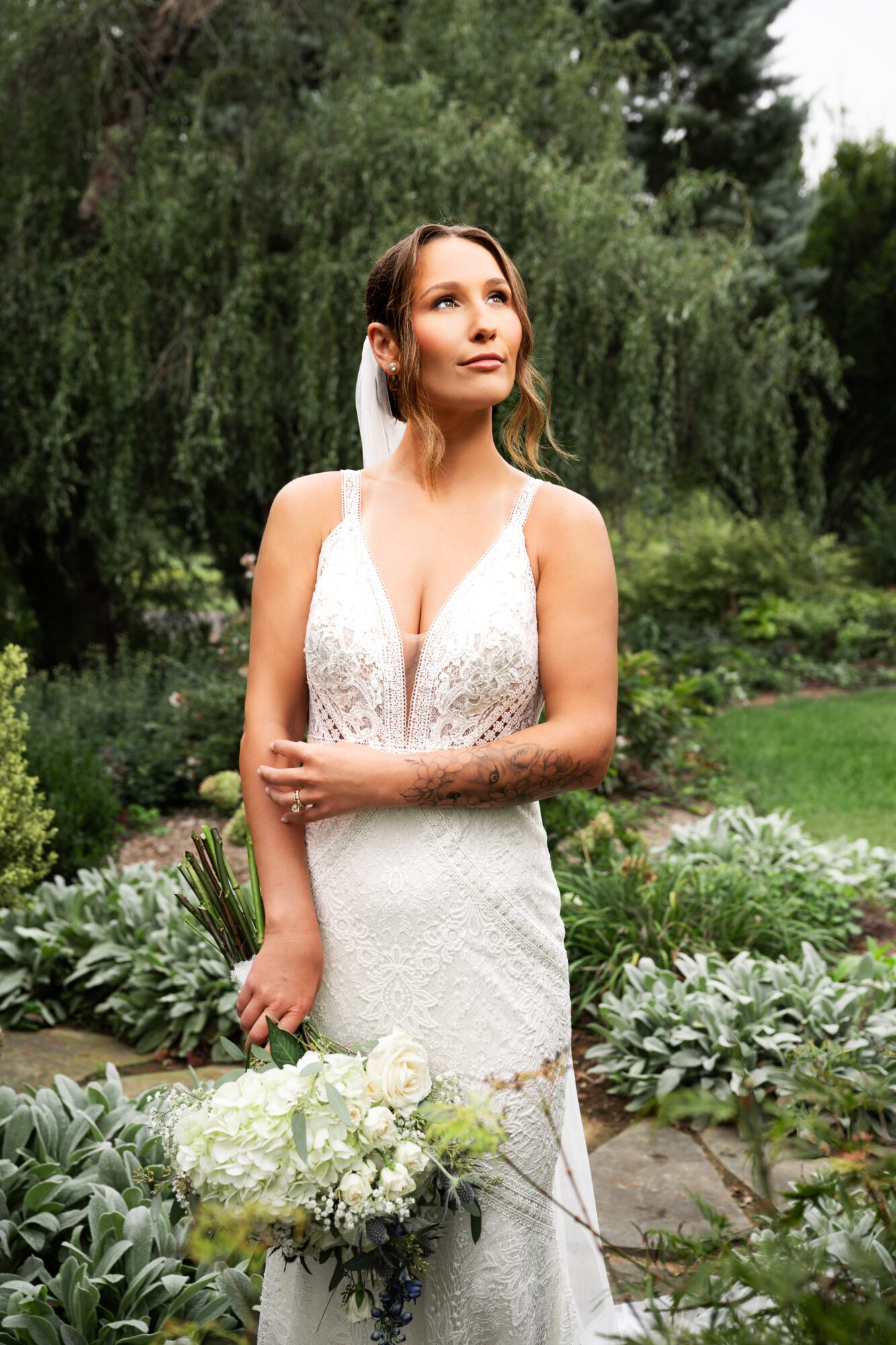 Woman in wedding dress holding bouquet outdoors with greenery and trees in background.