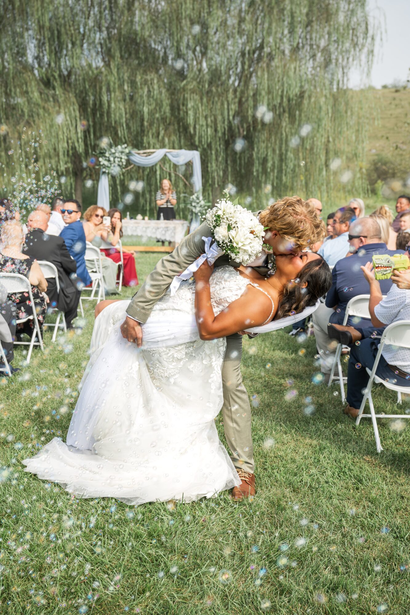 Bride and groom kissing outdoors, bride holding bouquet, guests seated nearby, wedding arch in background, grass and trees, bubbles floating.