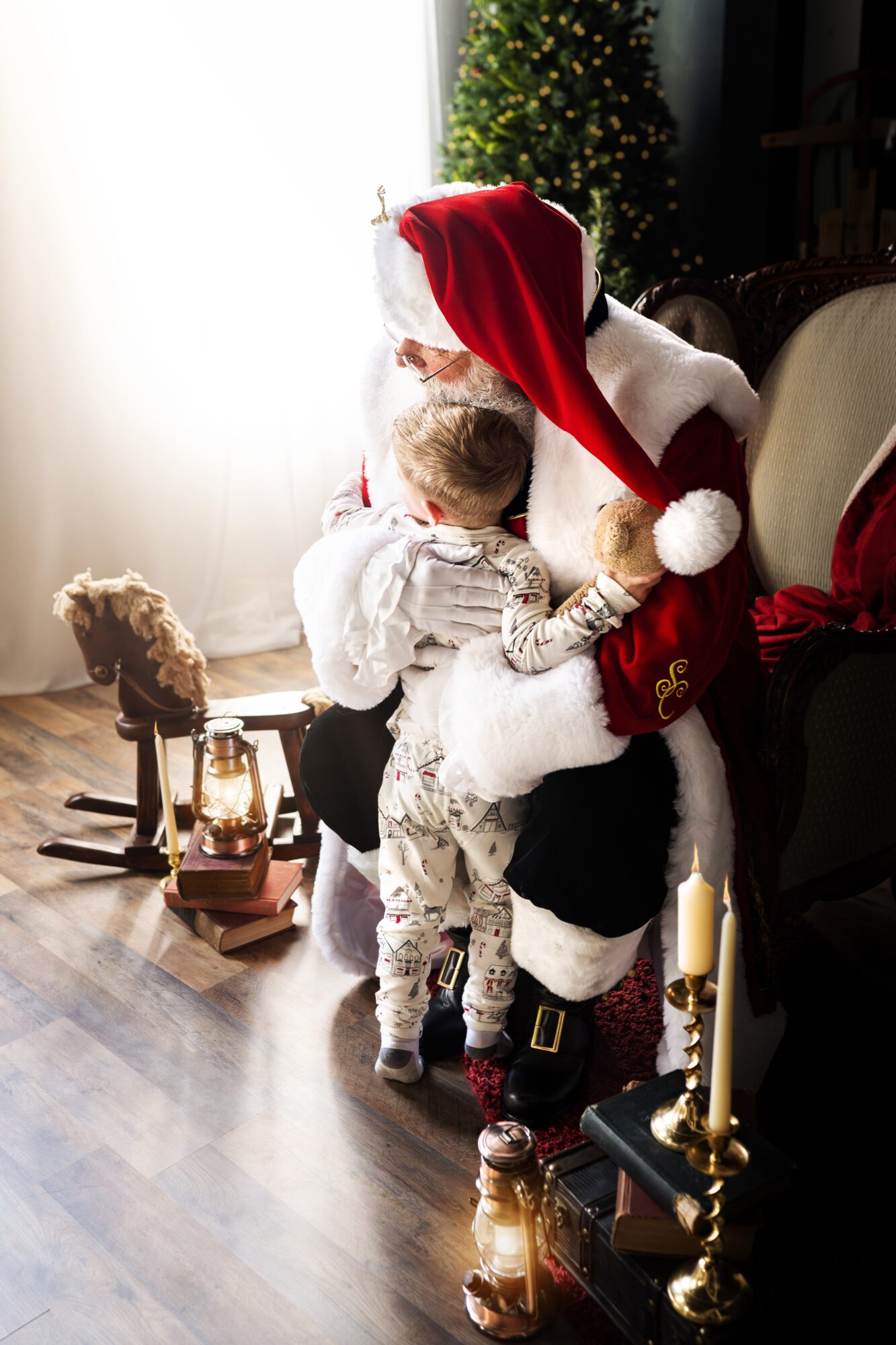 Child hugging a person dressed as Santa Claus with a Christmas tree in the background, near a window with curtains.