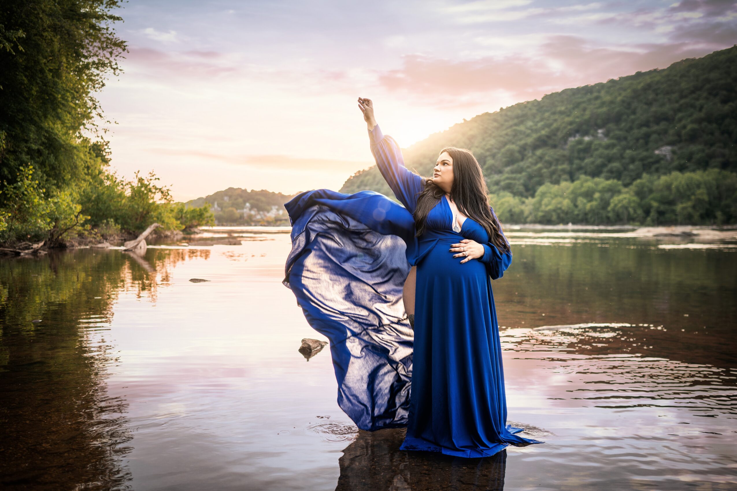 Pregnant woman in a blue dress standing in a river with her arm raised, surrounded by nature and a sunset sky.