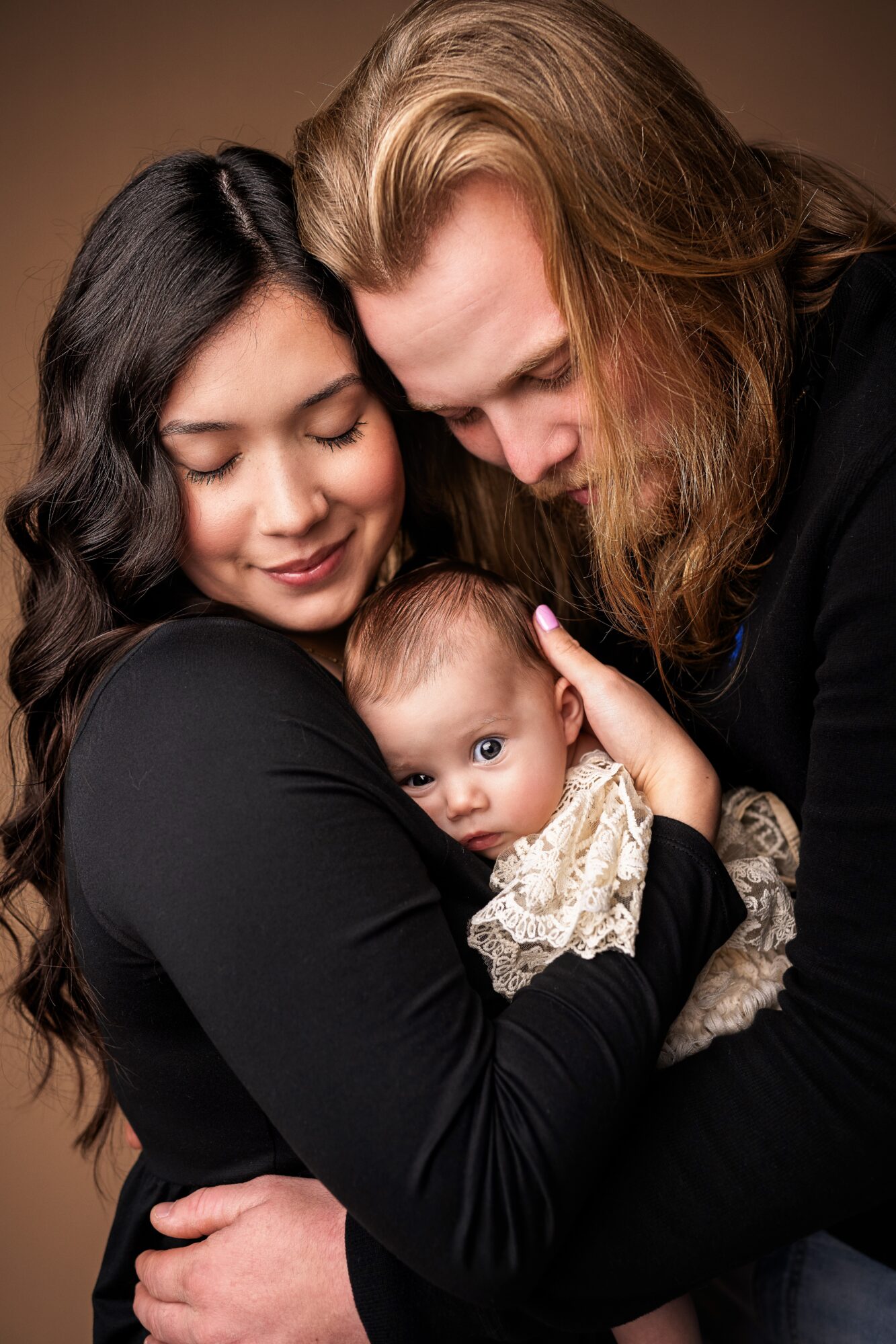 Family of three with a woman, man, and baby, close together, in warm lighting.