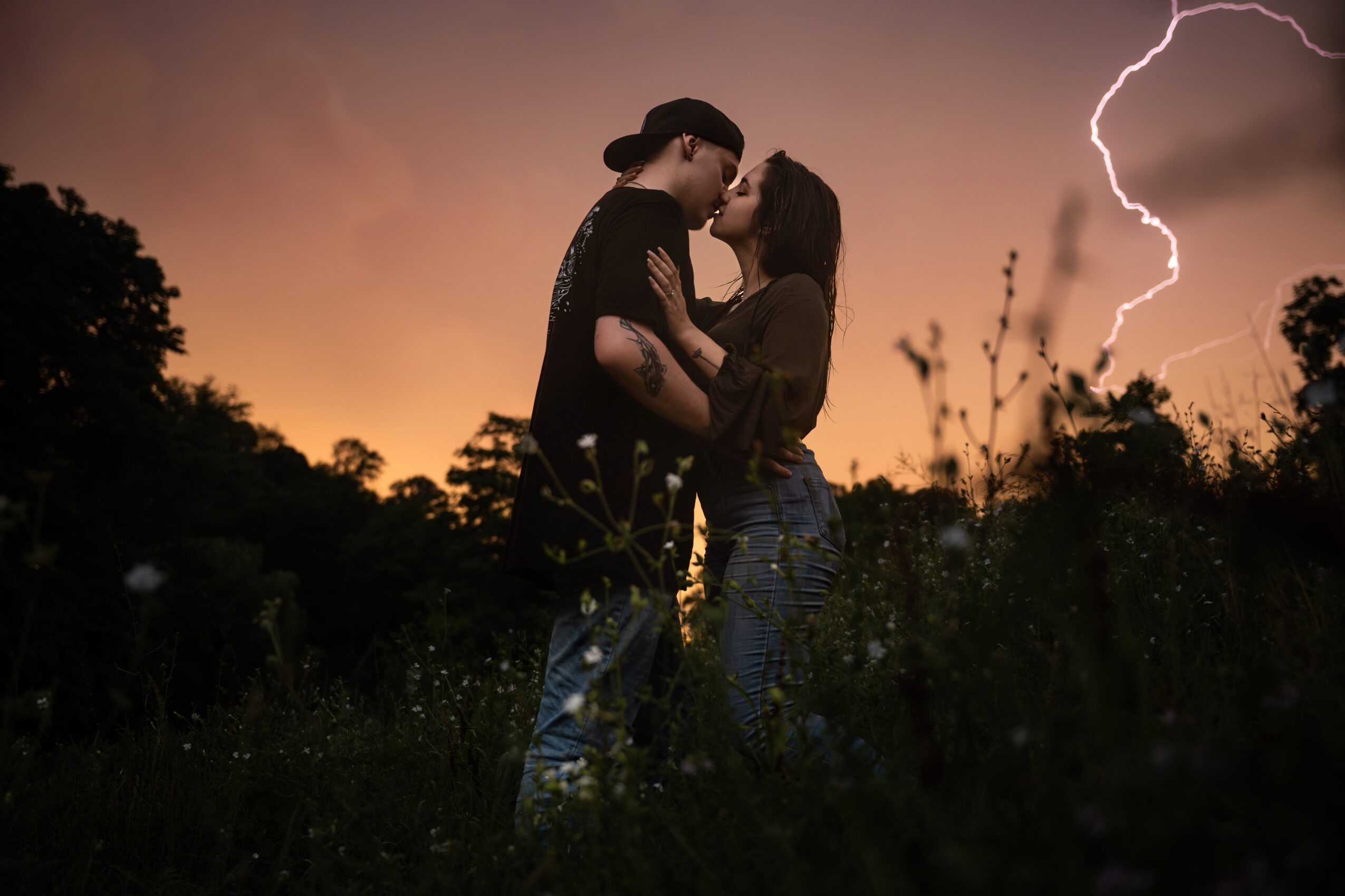 A couple stands close, embracing and kissing outdoors during a lightning storm at sunset, with trees and lightning in the background.