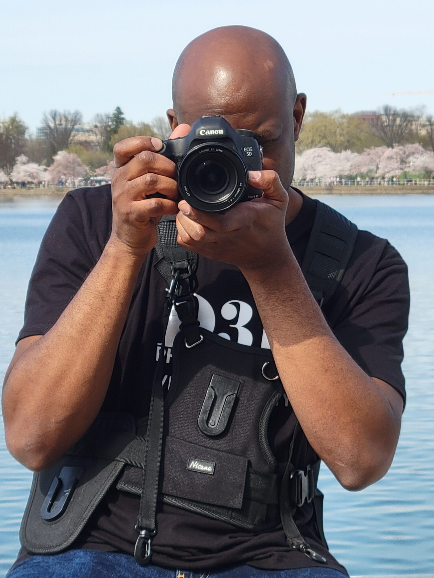 Person taking a photo with a camera near a body of water, with trees and landscape in the background.
