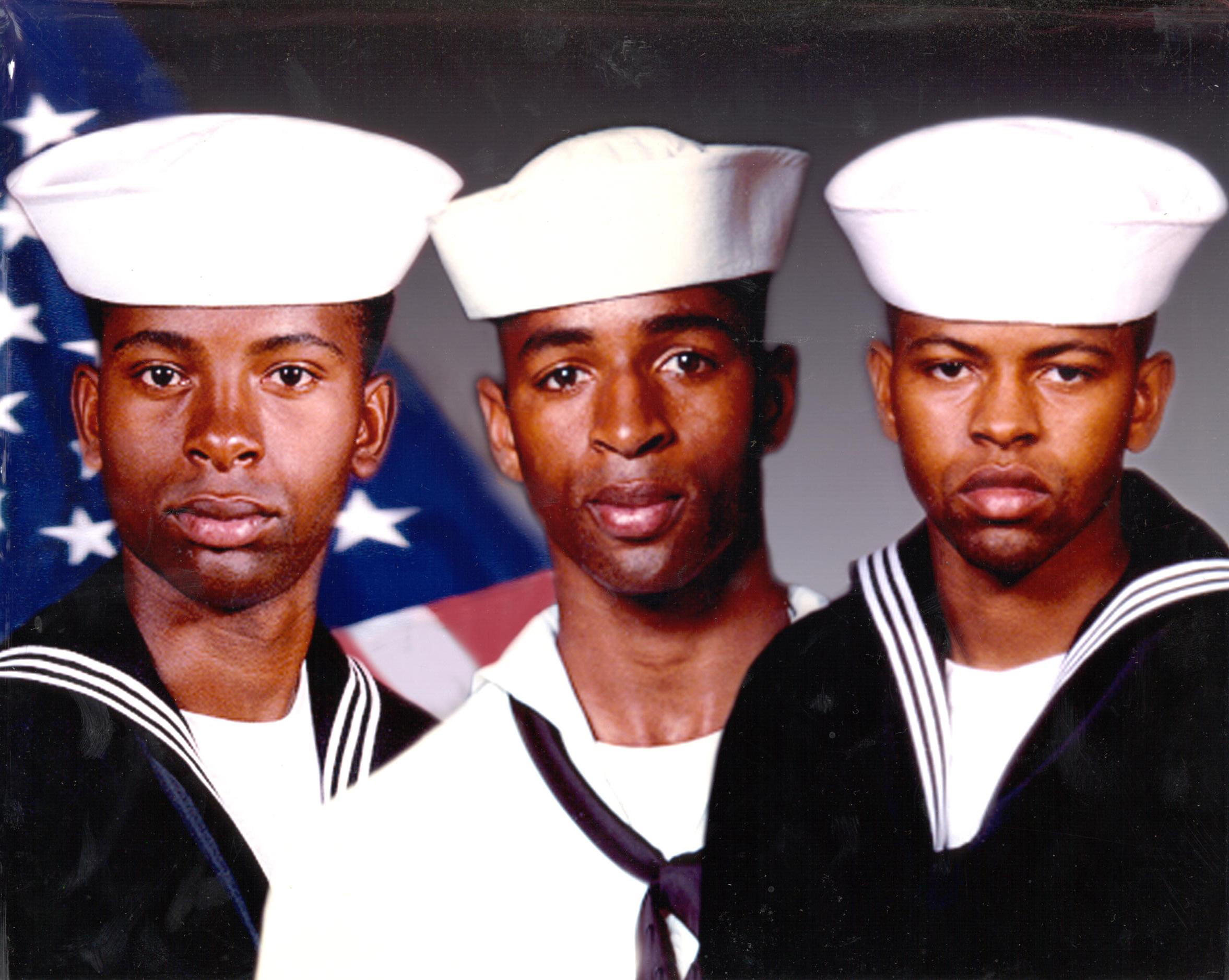 Three young men in sailor uniforms and white hats pose for a photo, with an American flag in the background.