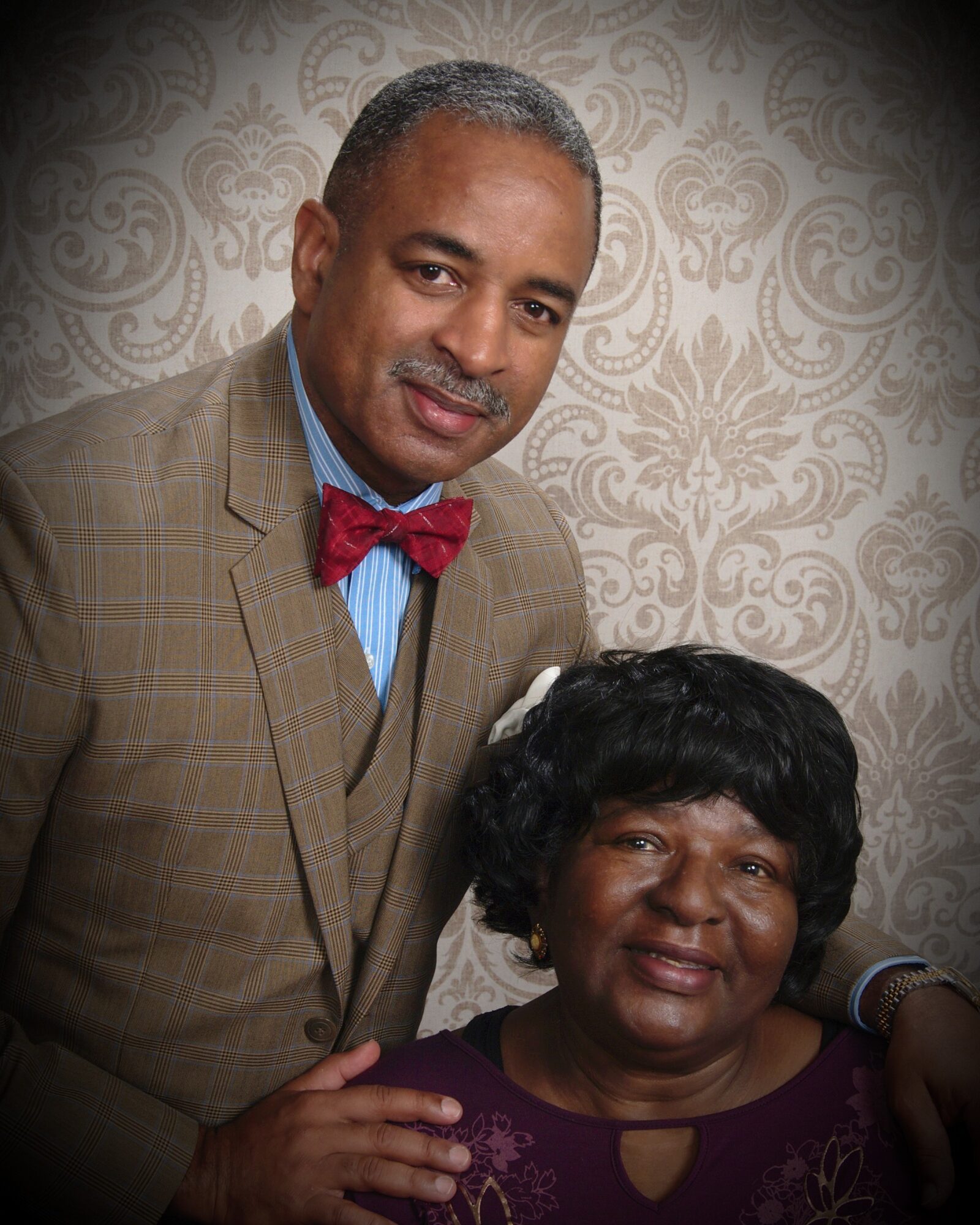 A man and woman pose together, the man wearing a suit with a red bow tie, the woman in a purple top, both smiling.