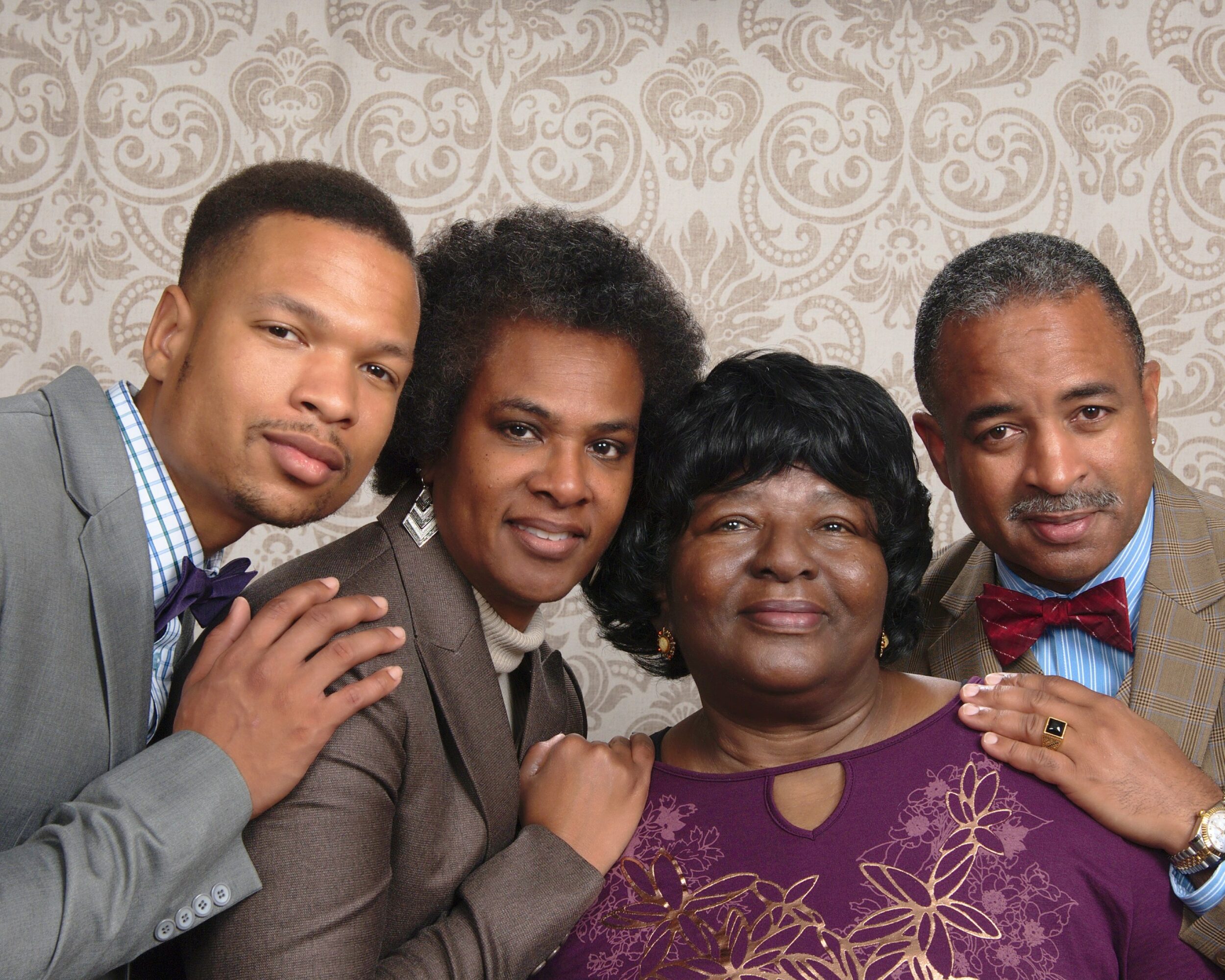 Four people standing close together, smiling, in front of a patterned wall, dressed in formal attire.