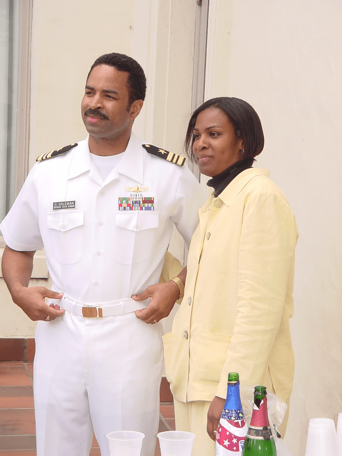 A man in a white military uniform and a woman in a yellow jacket stand together indoors, with drinks on a table nearby.