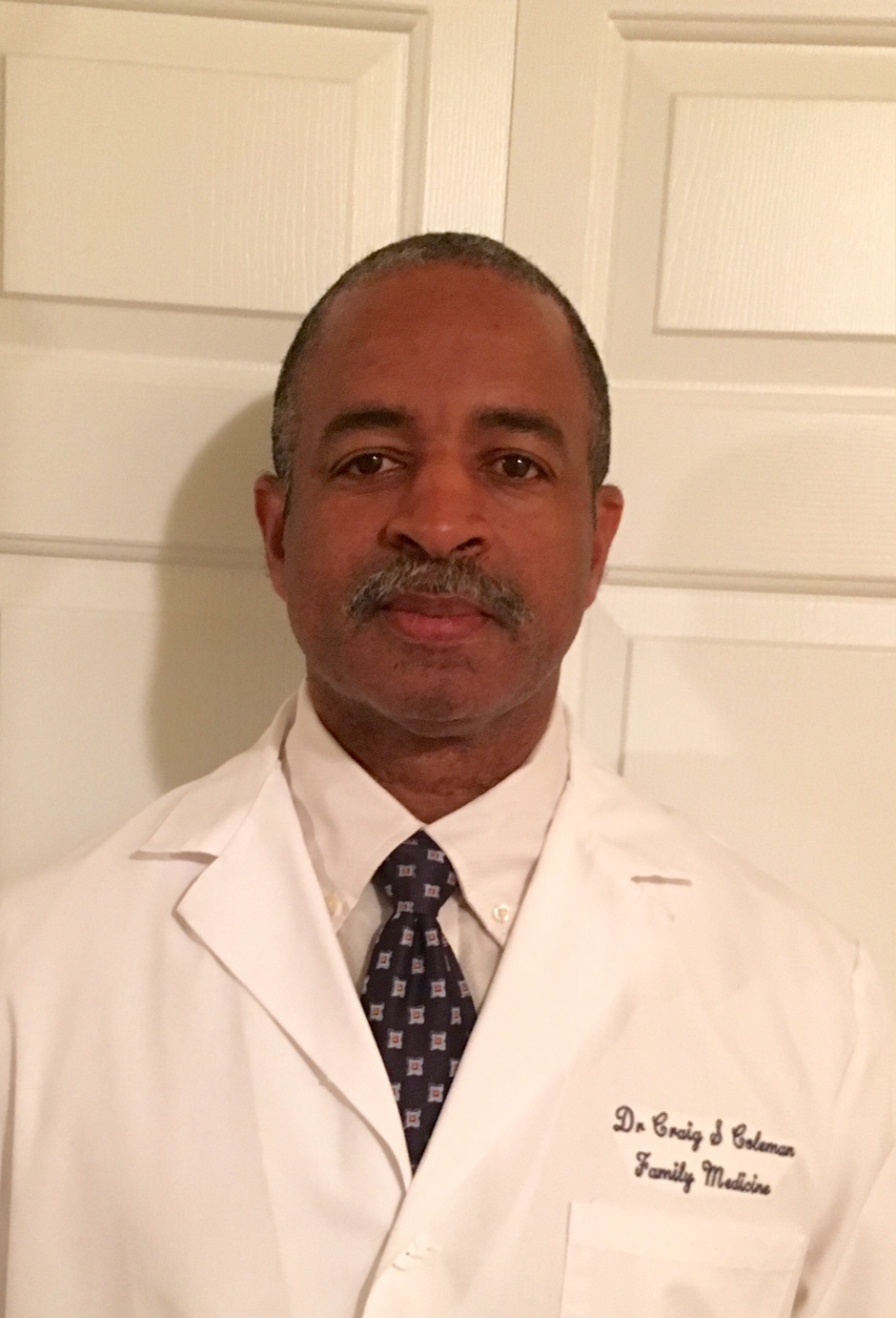 Man in white coat with tie, standing in front of white doors, looking at camera.