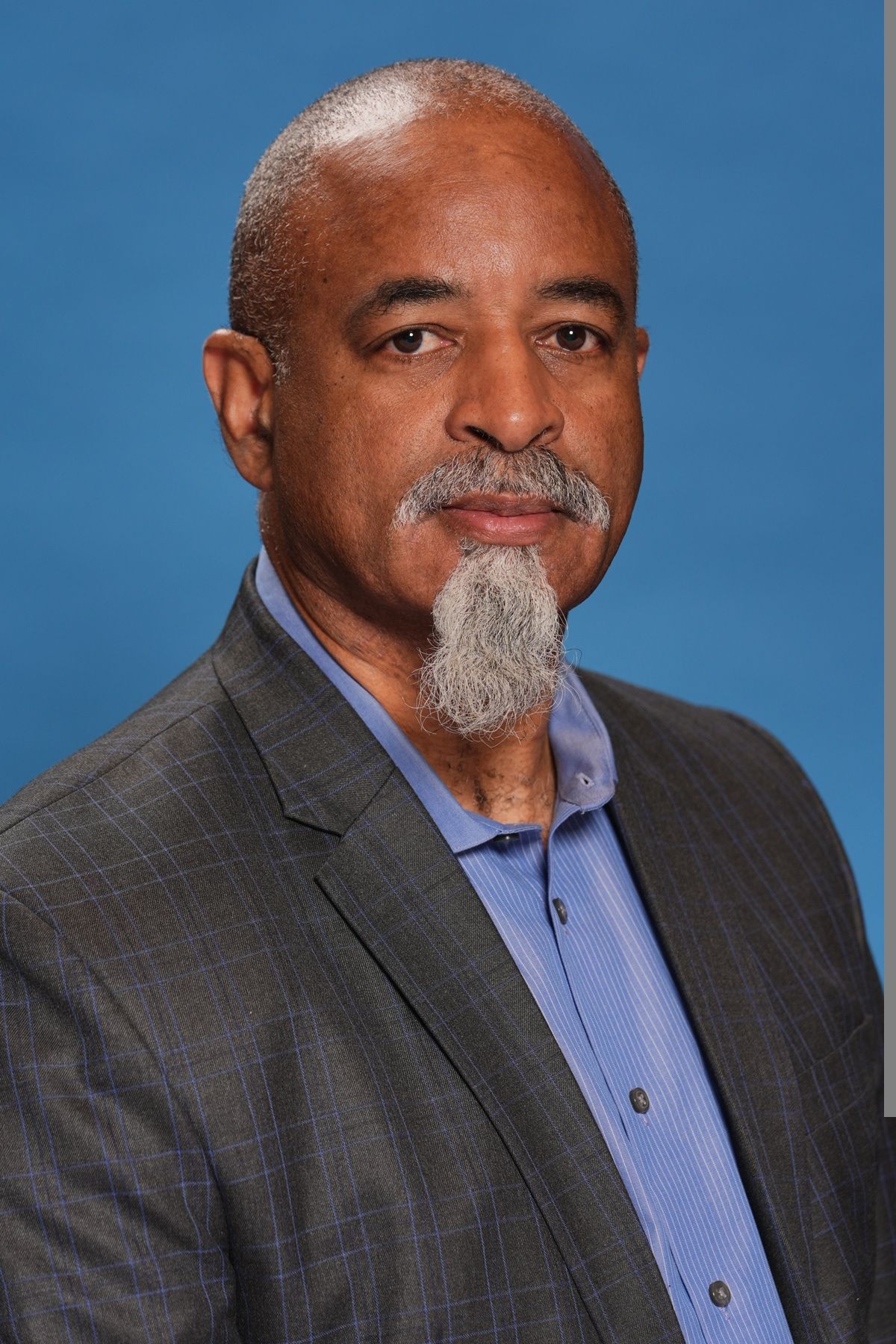 Portrait of an older man with gray hair and beard, wearing a suit and blue shirt, against a blue background.