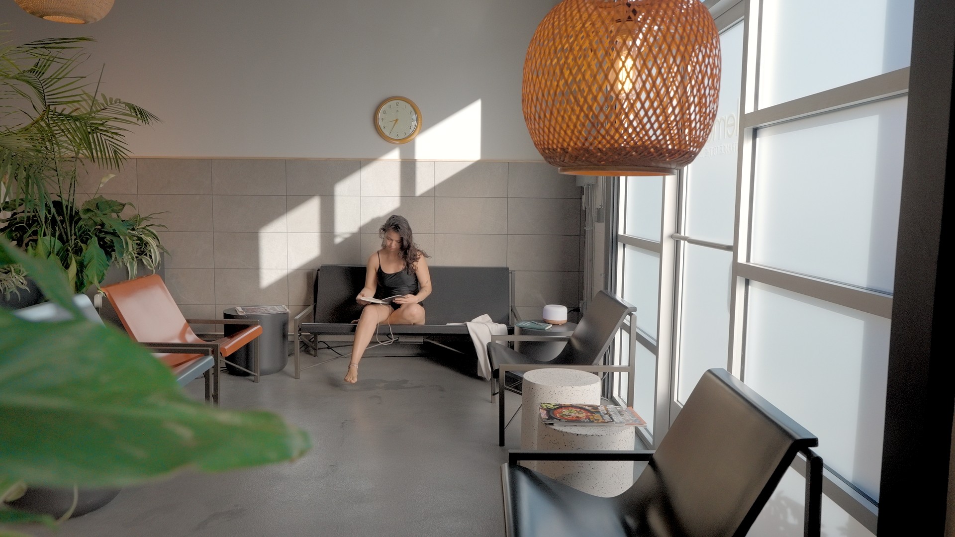 A woman sits on a gray sofa in a modern room with large windows, sunlight, and hanging light fixture.