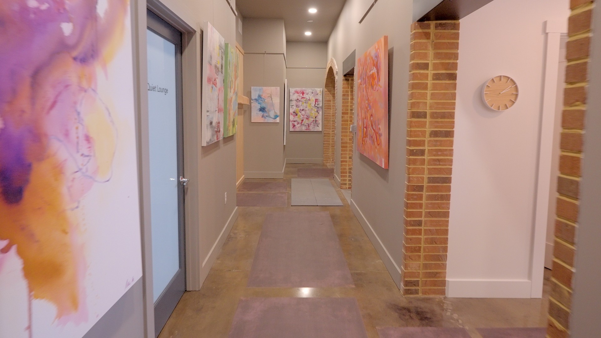 Hallway with artwork on walls, brick accents, and a door on the left, illuminated by ceiling lights.