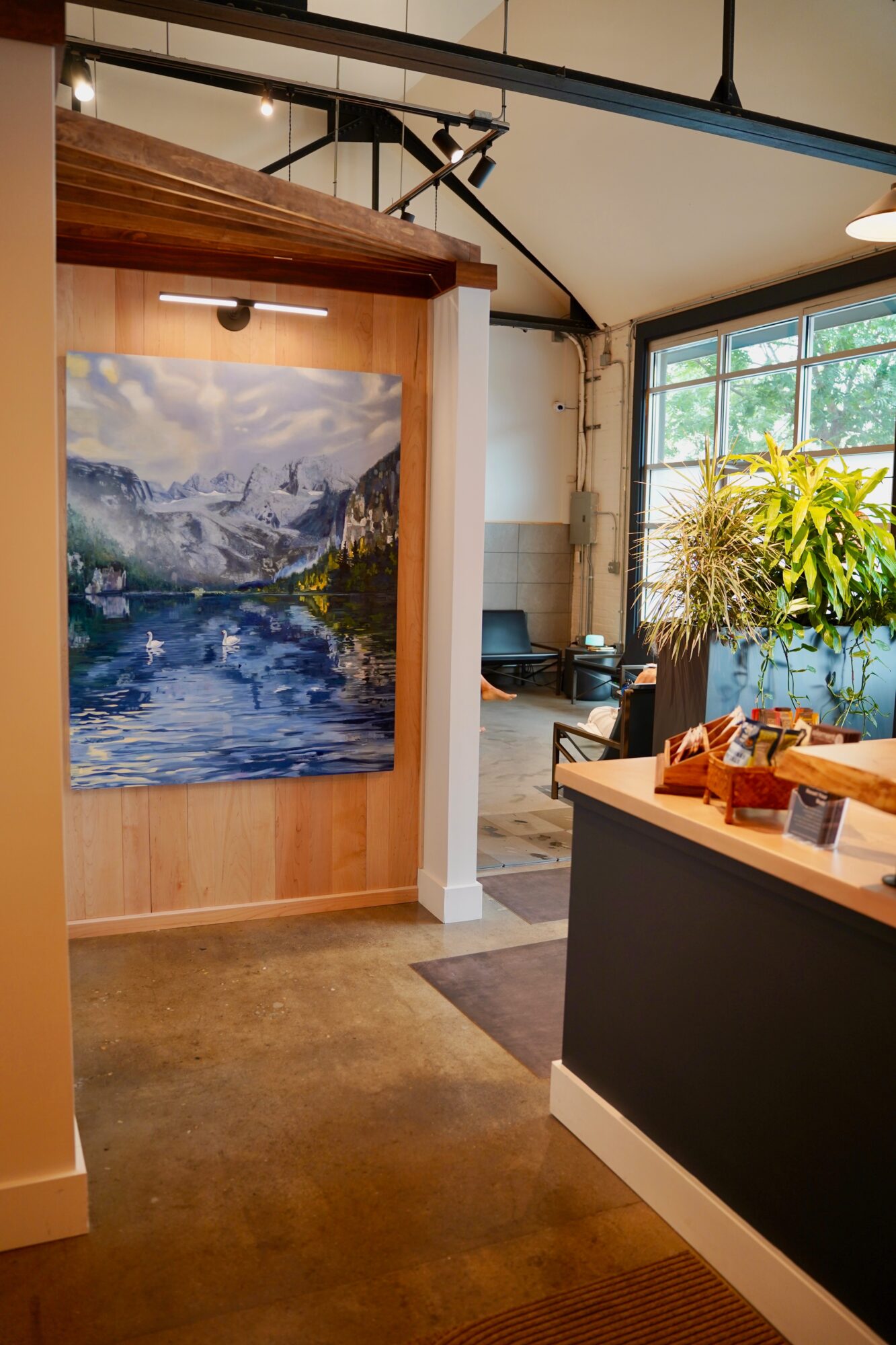 Interior of a cafe with a mountain and lake painting on the wall, large window with plants, and a counter with items.