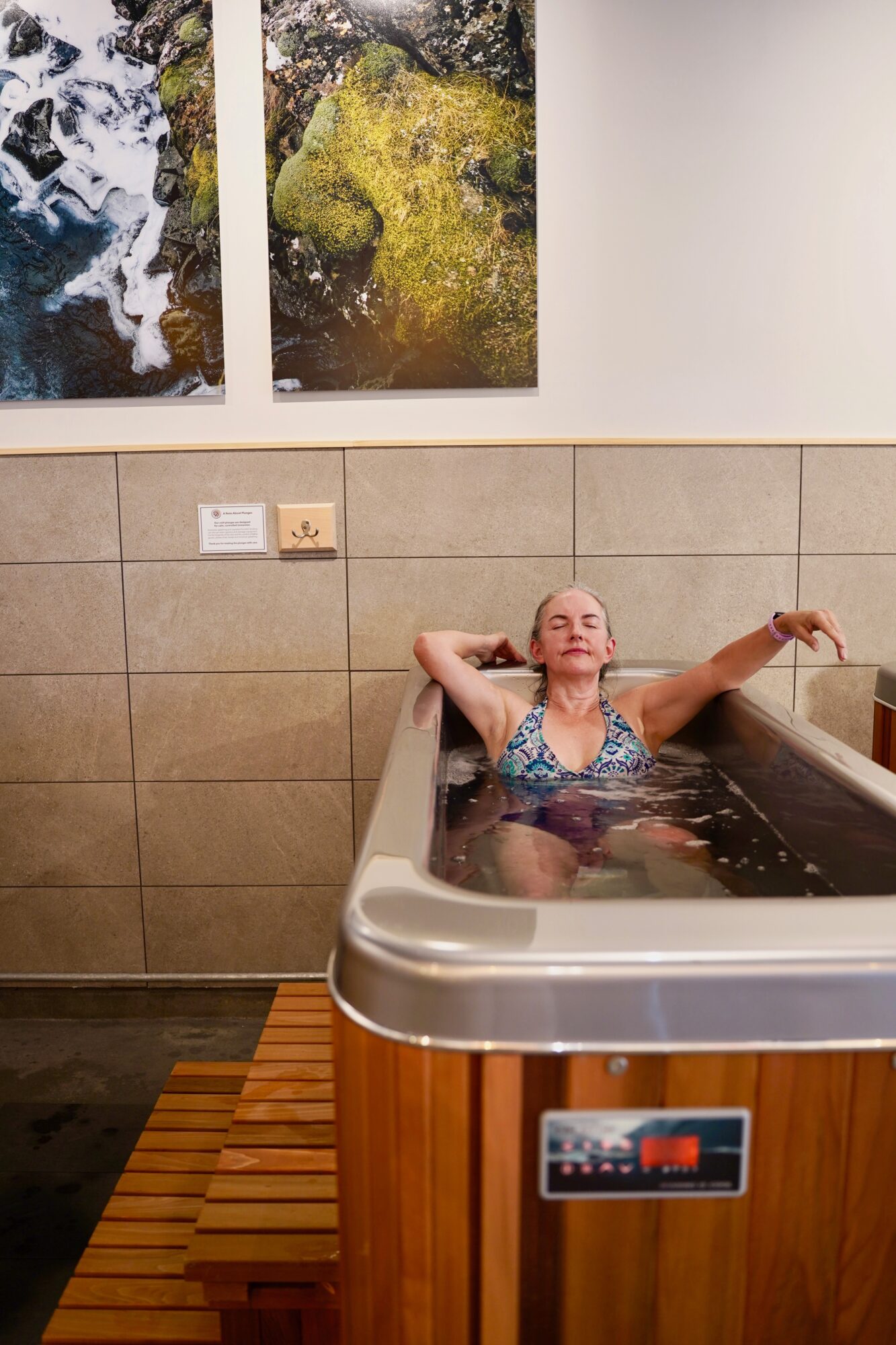 Woman relaxing in a hot tub with her eyes closed, arms stretched out, in a room with tiled walls and nature photos above.