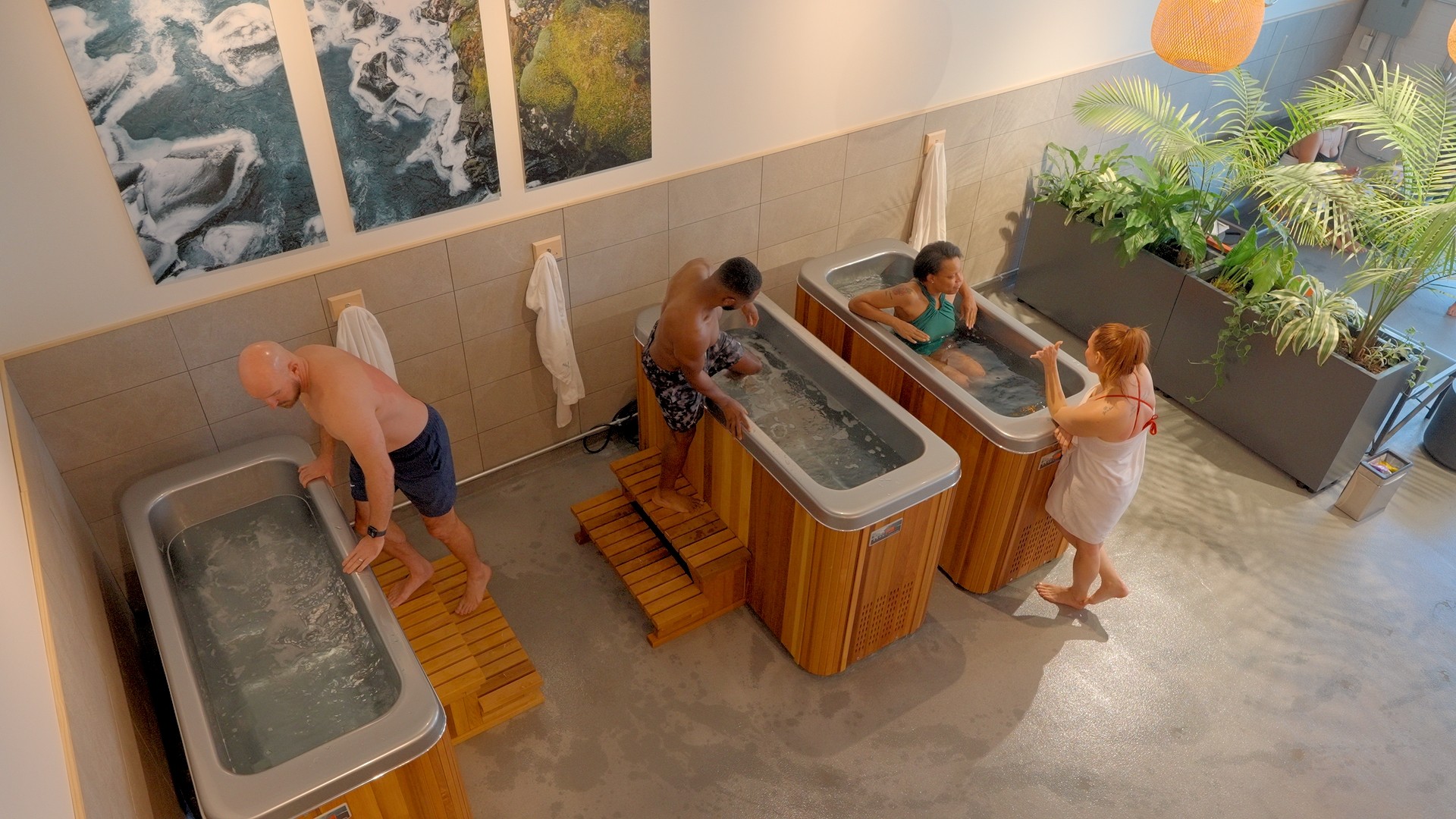Three people using foot baths with wooden platforms in a room with large plants and artwork on the wall.